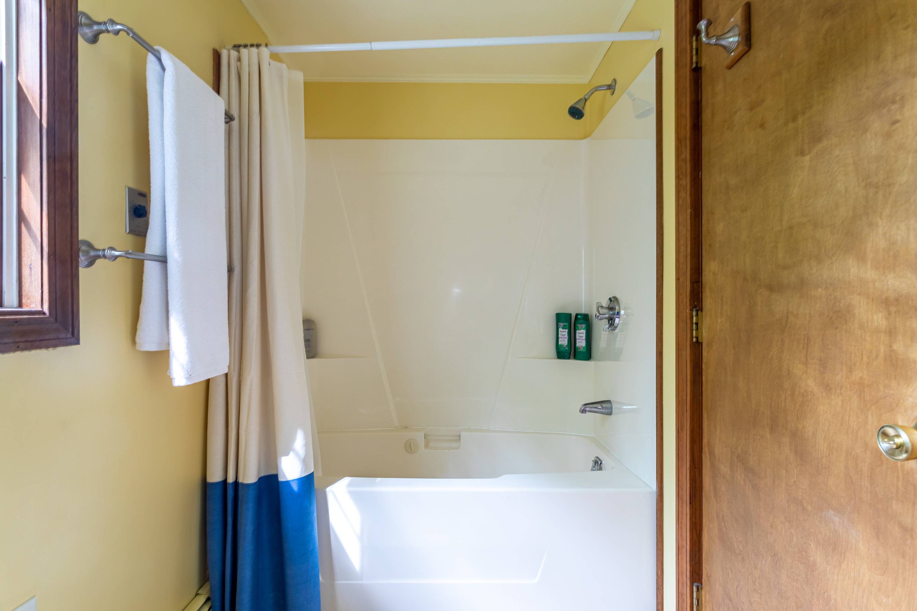 White shower-over-tub combo with a cream and blue shower curtain, featuring a wall-mounted showerhead and a small shelf with toiletries.