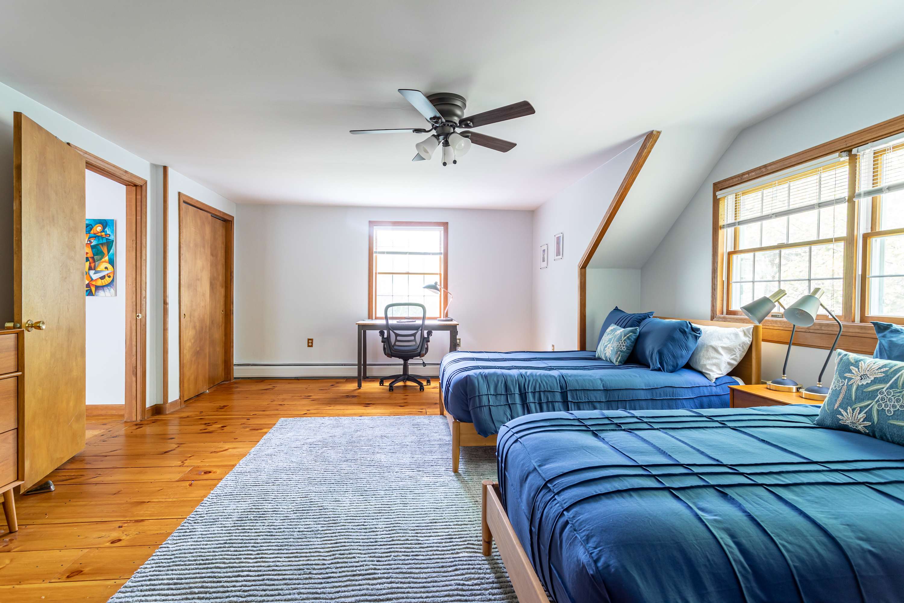 Spacious bedroom with two twin beds featuring blue bedding, light wood floors, a desk with a chair, and a ceiling fan, with natural light from a large window.