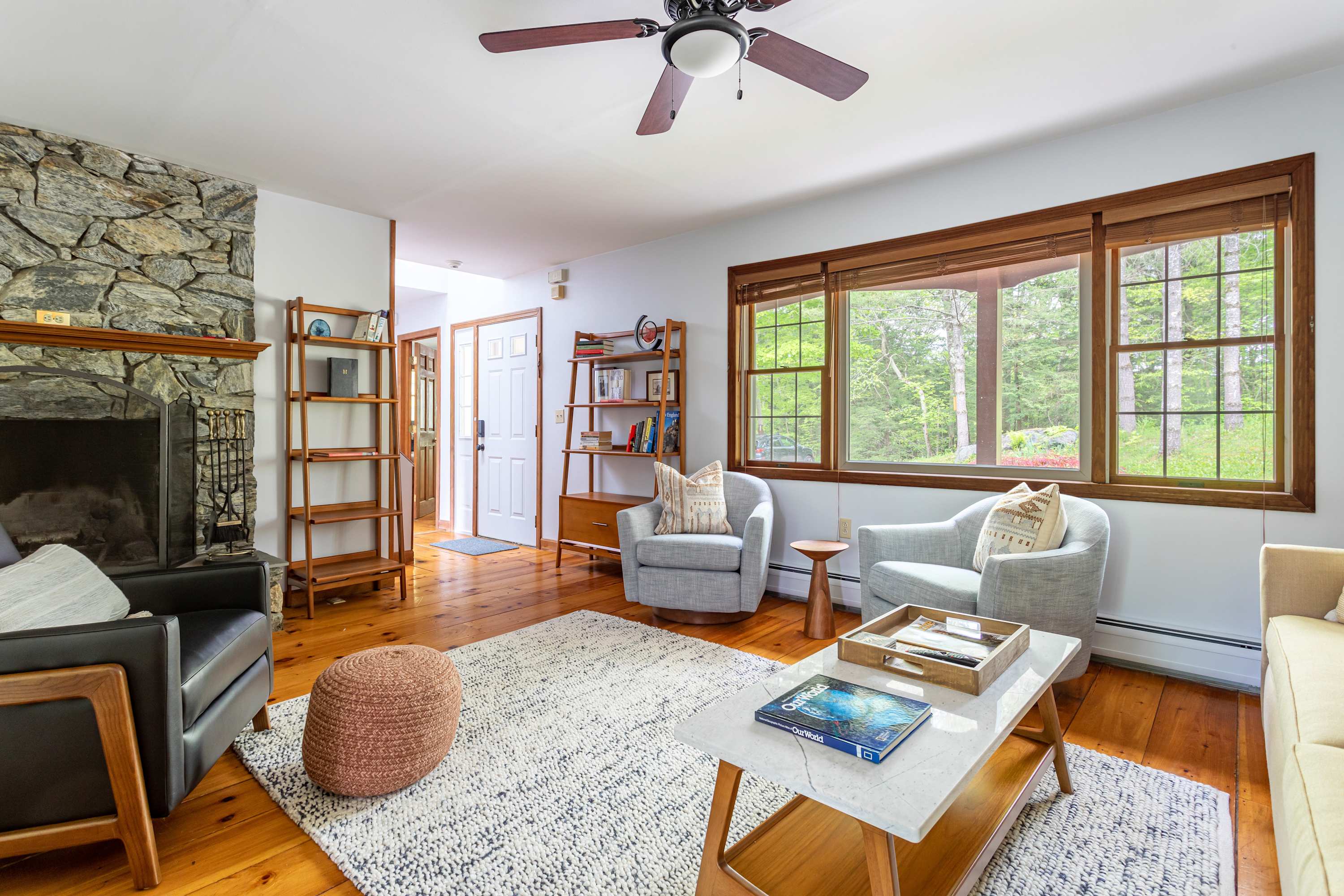 A bright living room with warm wooden floors and white walls features a stone fireplace, two light gray swivel chairs, a black leather armchair, and a large window overlooking the surrounding greenery.