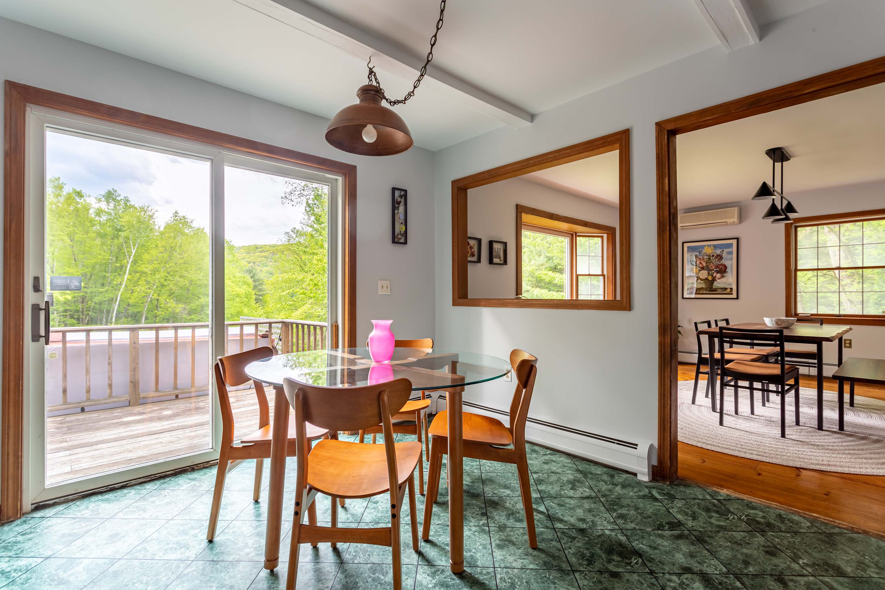 An inviting dining area with green tiled floors, a glass-top table, and wooden chairs, positioned next to a large sliding glass door opening to a deck with scenic outdoor views. A large wooden-framed mirror reflects an adjacent room, adding depth to the space.