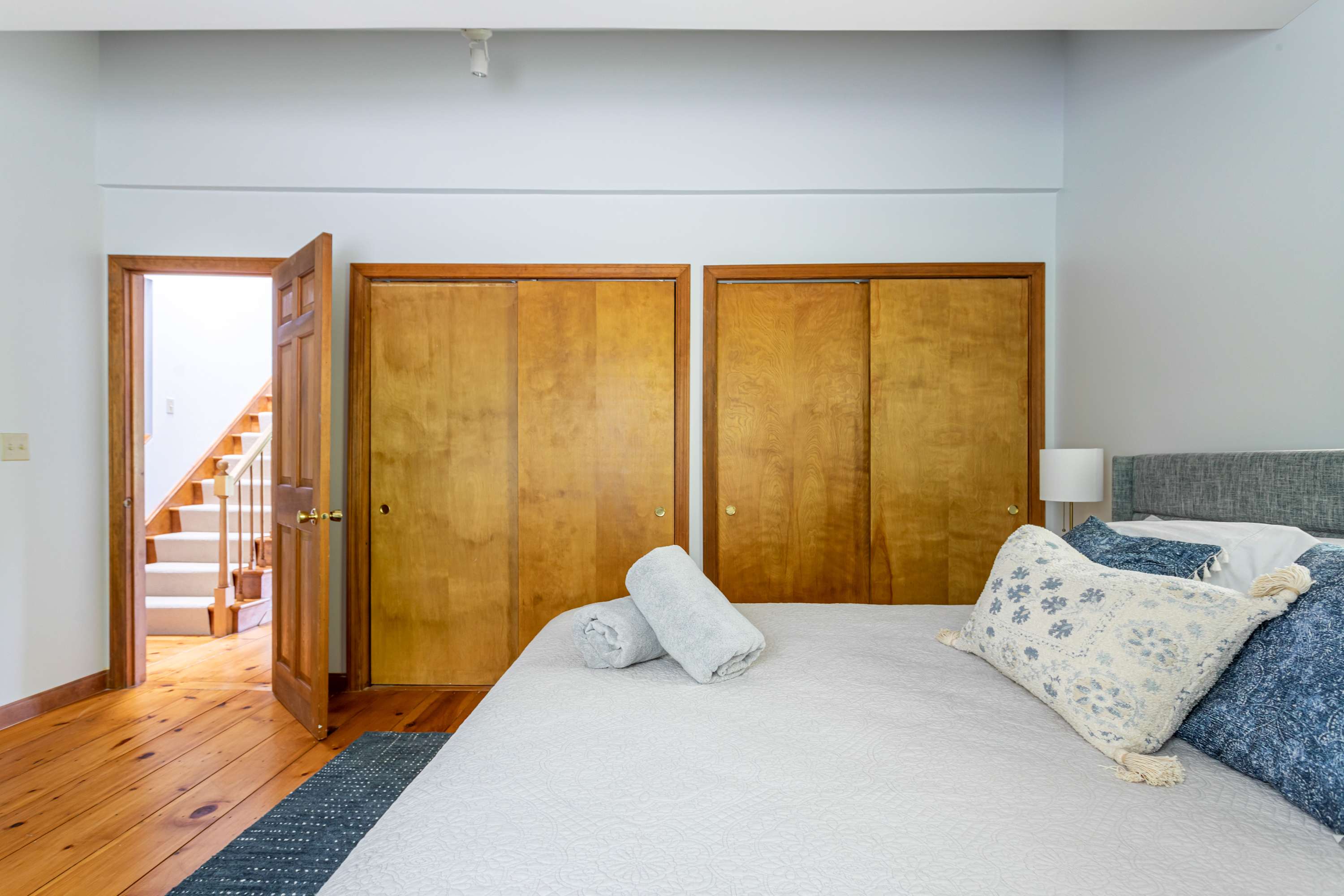 An interior view of bedroom-1, highlighting the entrance doorway and two wooden sliding closet doors. The room features light wood floors and light blue walls, with a glimpse of the bed and a nightstand, and a staircase visible through the open door.