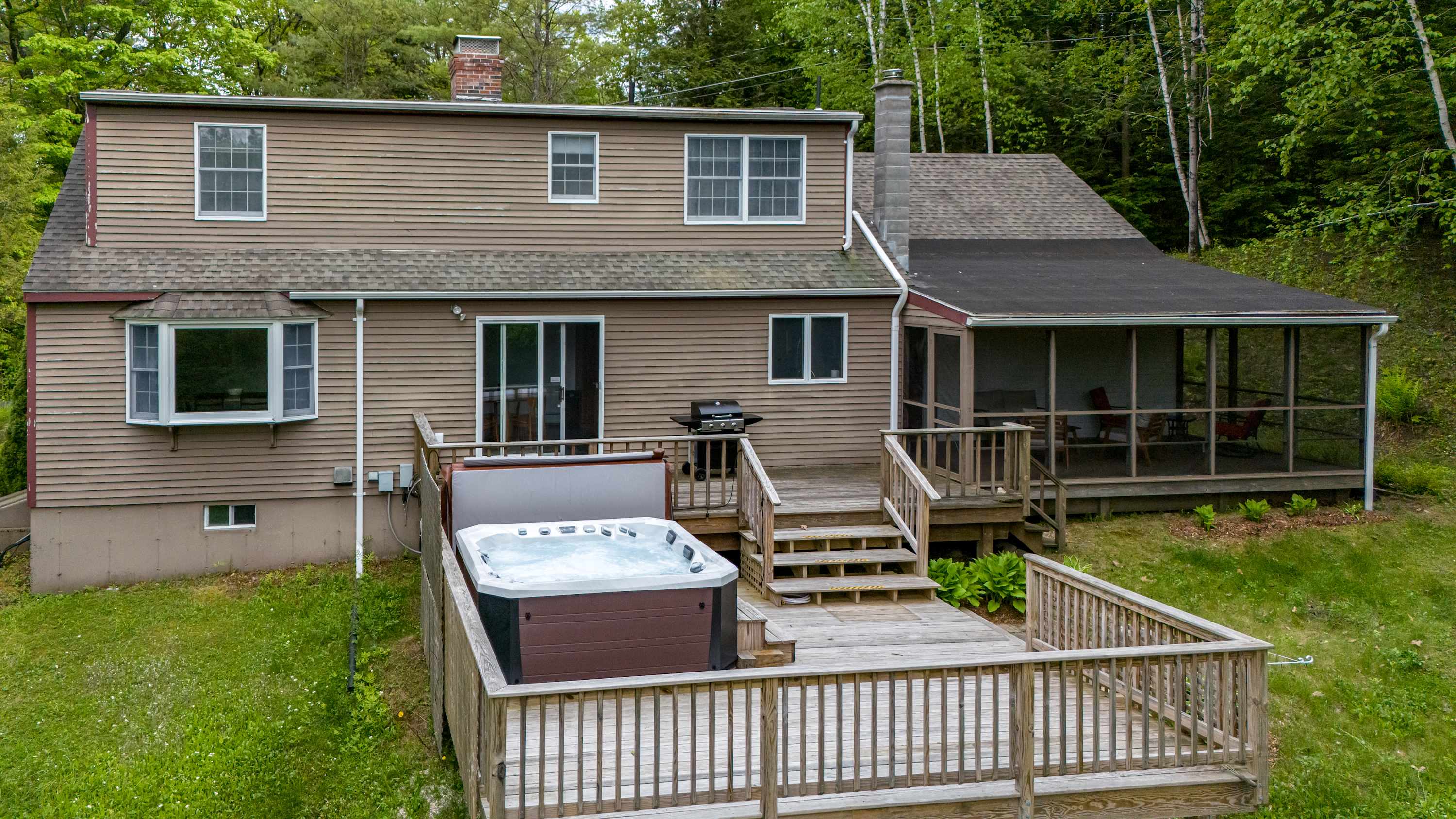 Rear exterior view of the house with light brown siding, featuring a multi-level wooden deck with a hot tub, a BBQ grill, and a screened porch.