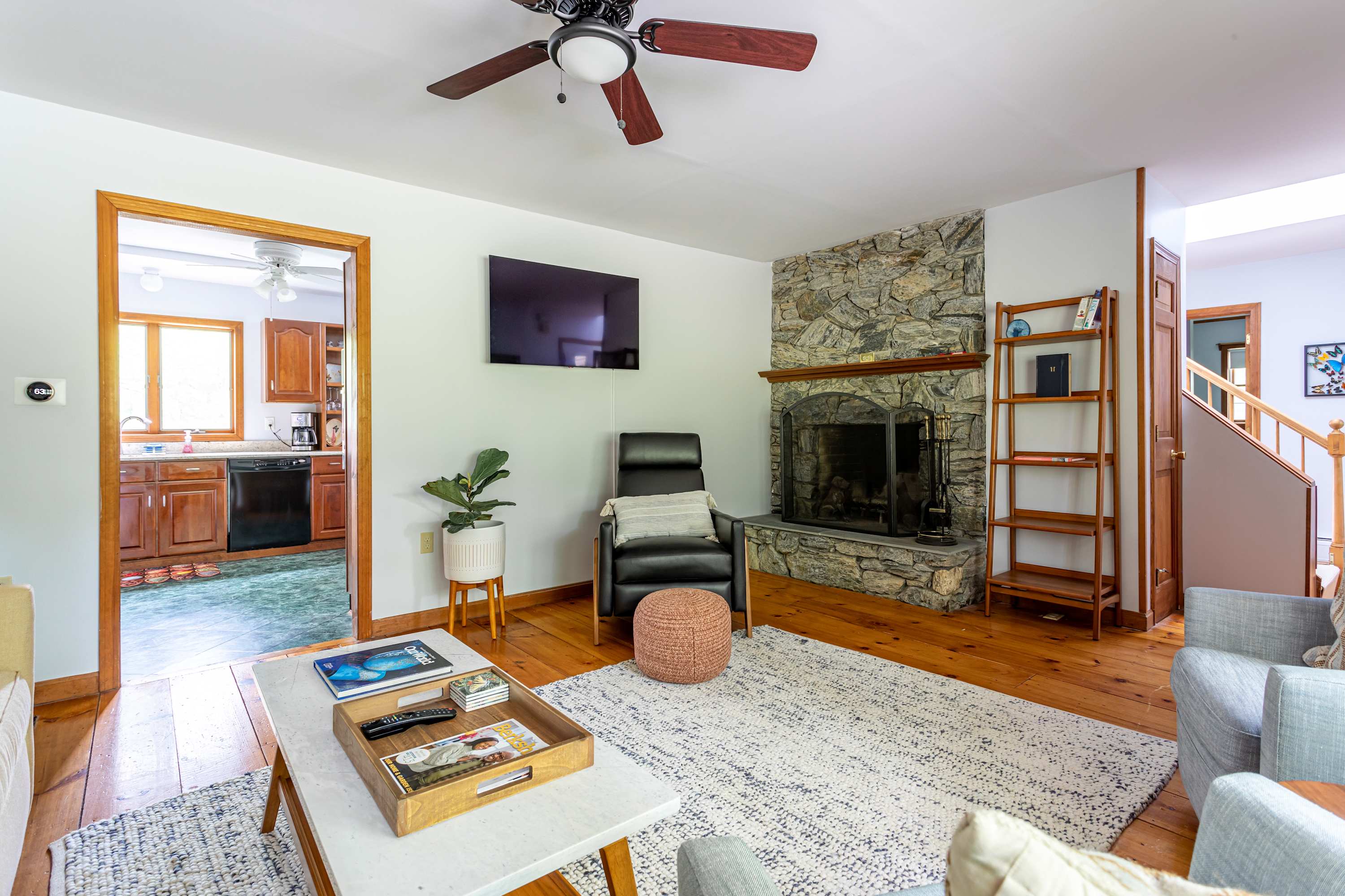 A cozy living room with warm wooden floors and white walls features a prominent stone fireplace, a wall-mounted TV, a black leather armchair, and a ceiling fan, with a view into the kitchen.