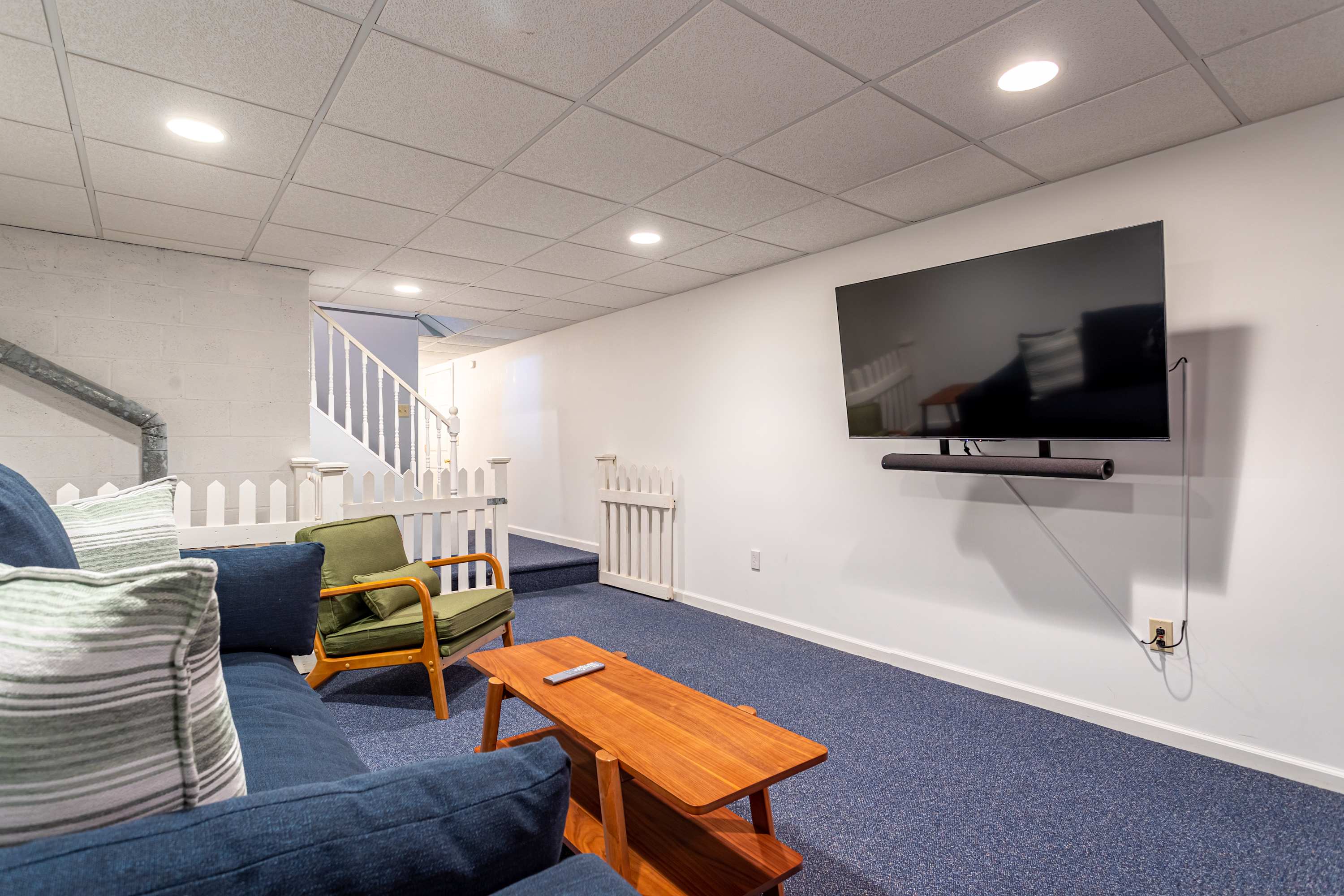 Cozy media room with a large wall-mounted TV, a blue sofa, a wooden coffee table, and blue carpet, featuring a white picket fence railing leading to stairs.