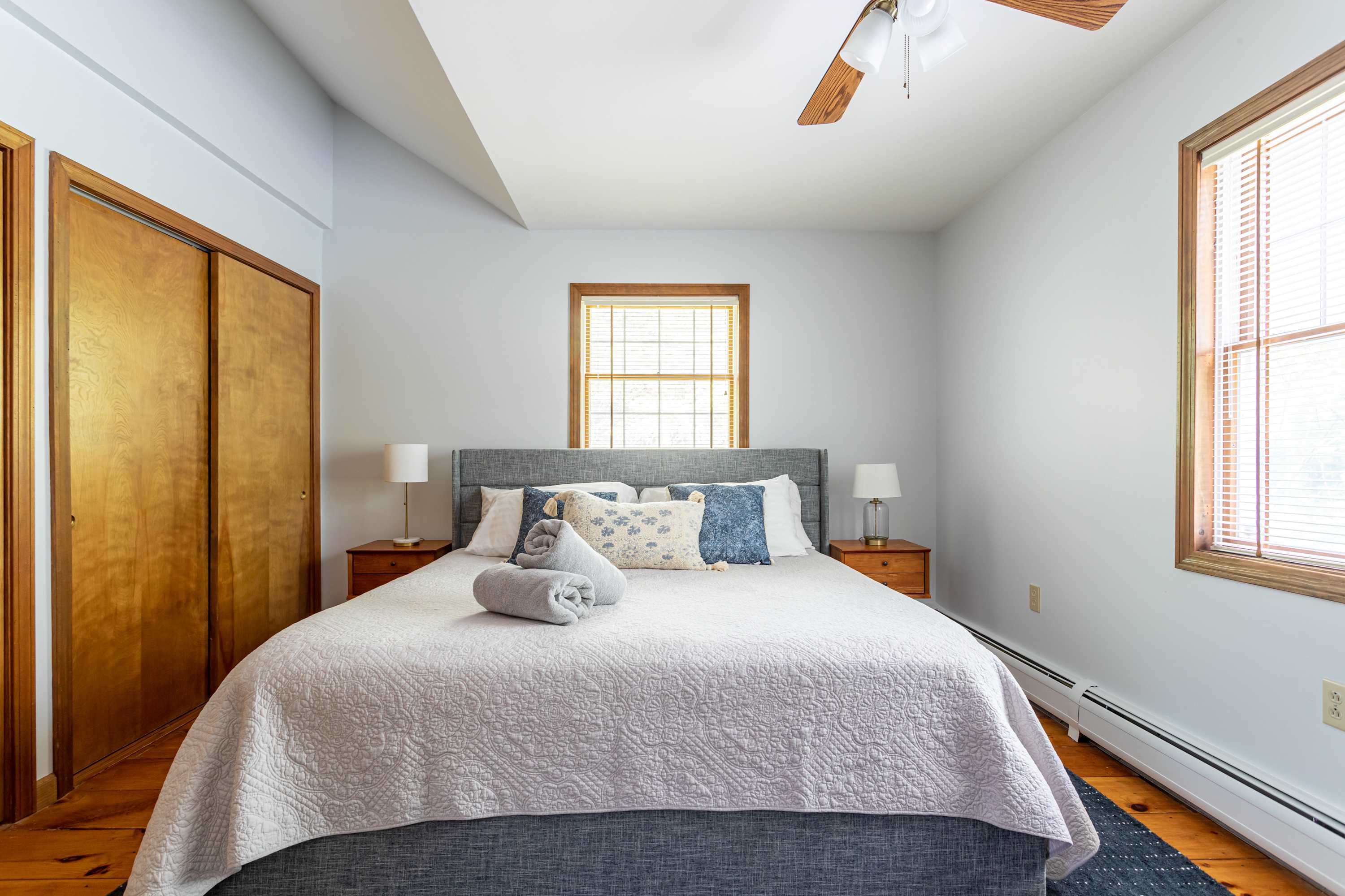 A serene bedroom featuring light blue walls, a queen-sized bed with a gray upholstered headboard, and light wood floors. Two wooden closets with sliding doors are visible, and a window with blinds offers natural light, creating a peaceful retreat.