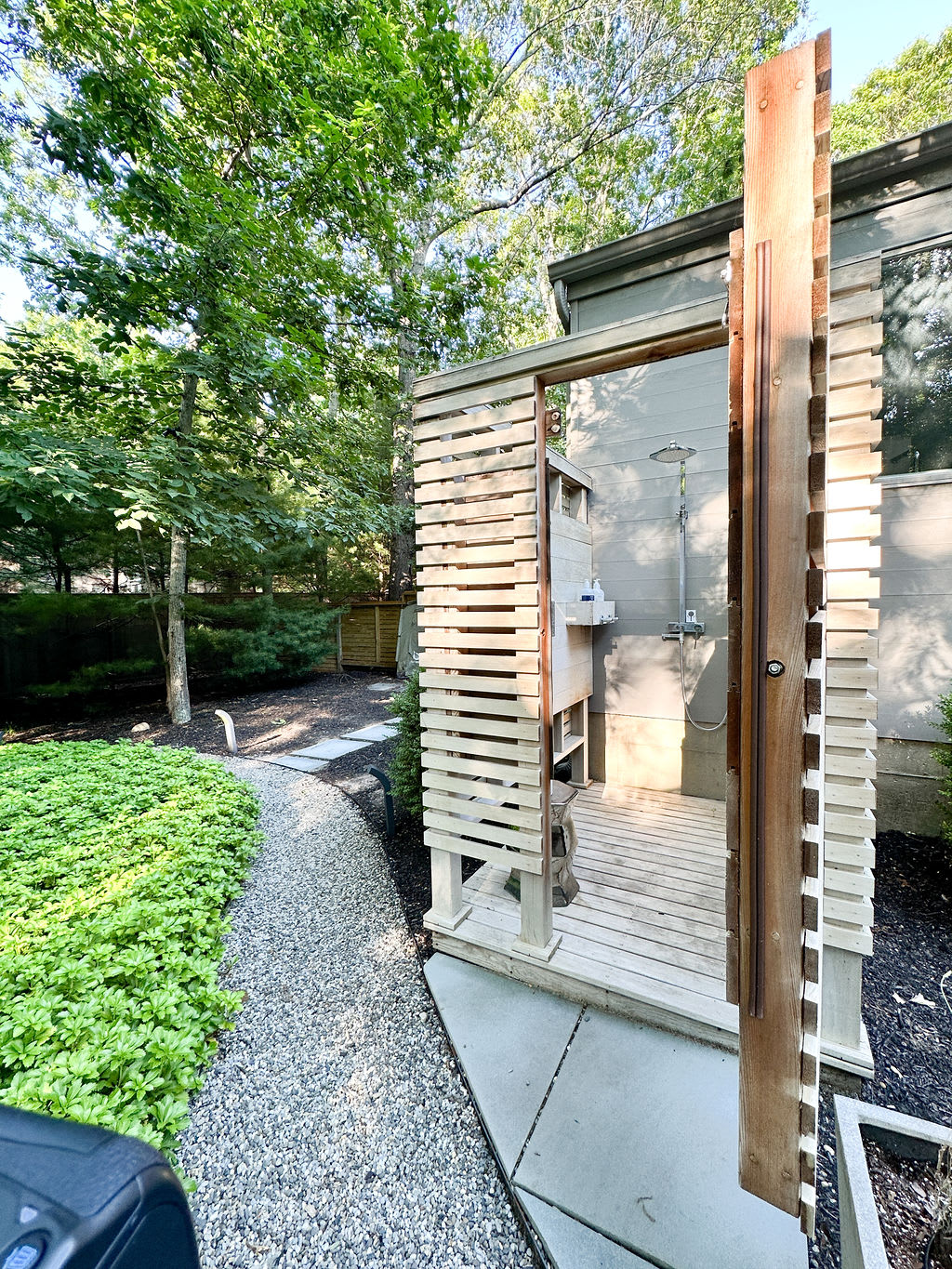 Modern outdoor shower enclosure with slatted wooden walls and a concrete base, featuring a rain showerhead and a small shelf. A gravel path and lush green ground cover are visible in the surrounding yard.