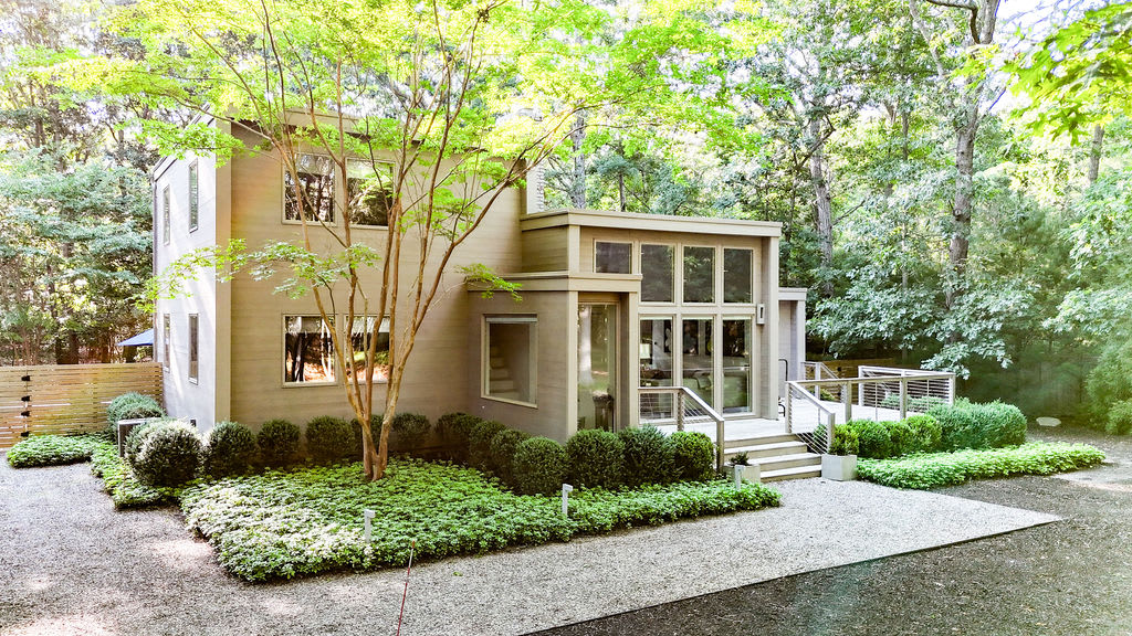 Contemporary two-story house with light gray siding and large windows, surrounded by meticulously landscaped greenery and a gravel driveway. A mature tree with vibrant green leaves stands prominently in the front yard.