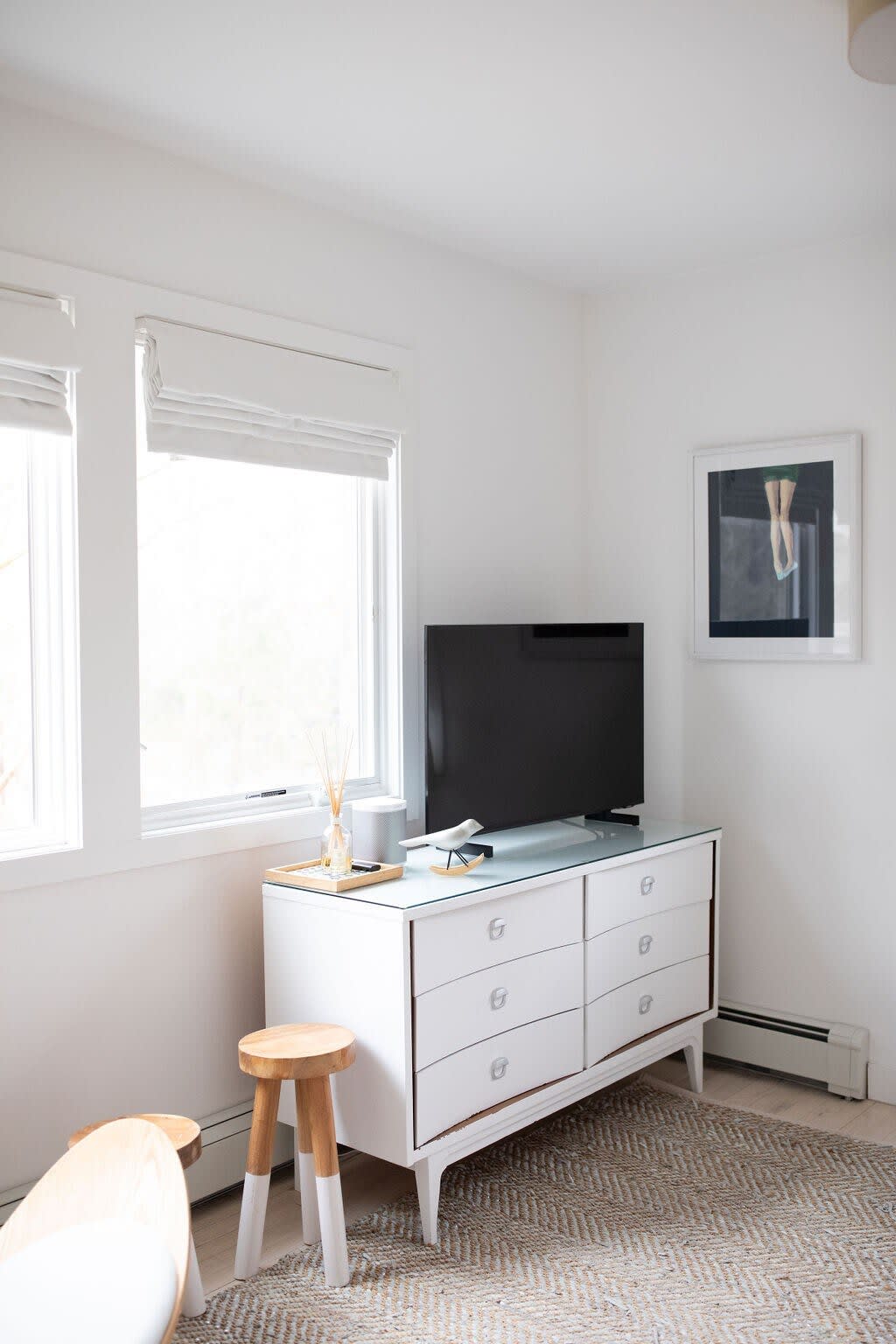 Bright bedroom featuring a white dresser with a TV, a patterned rug, and two windows with white blinds, creating a clean and minimalist aesthetic.