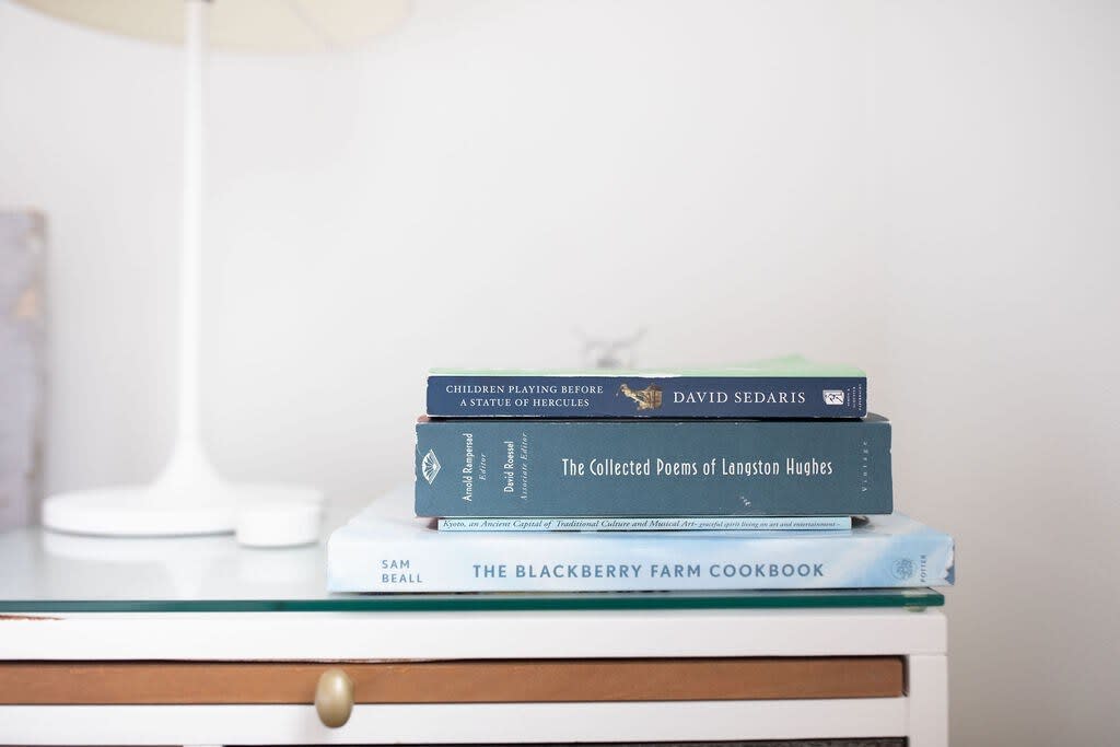 Stack of three books, including "Children Playing Before a Statue of Hercules" and "The Collected Poems of Langston Hughes," resting on a glass-topped bedside table.