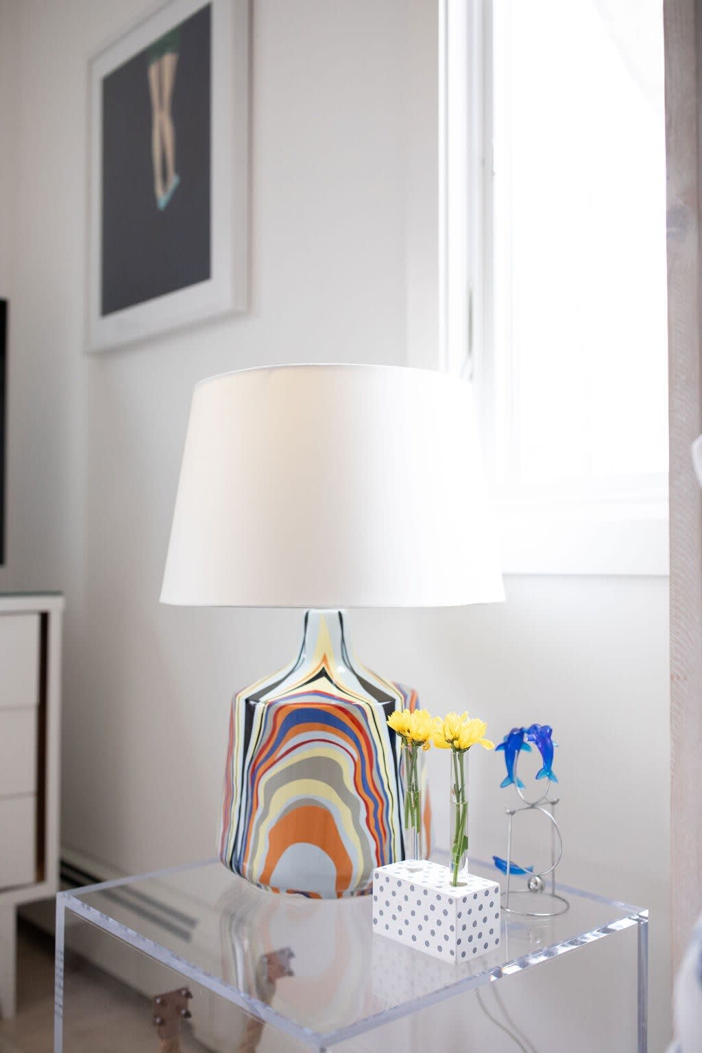 Close-up of a clear acrylic bedside table with a vibrant, multi-colored lamp, a small vase with yellow flowers, and blue dolphin figurines, adding a pop of color to the room.
