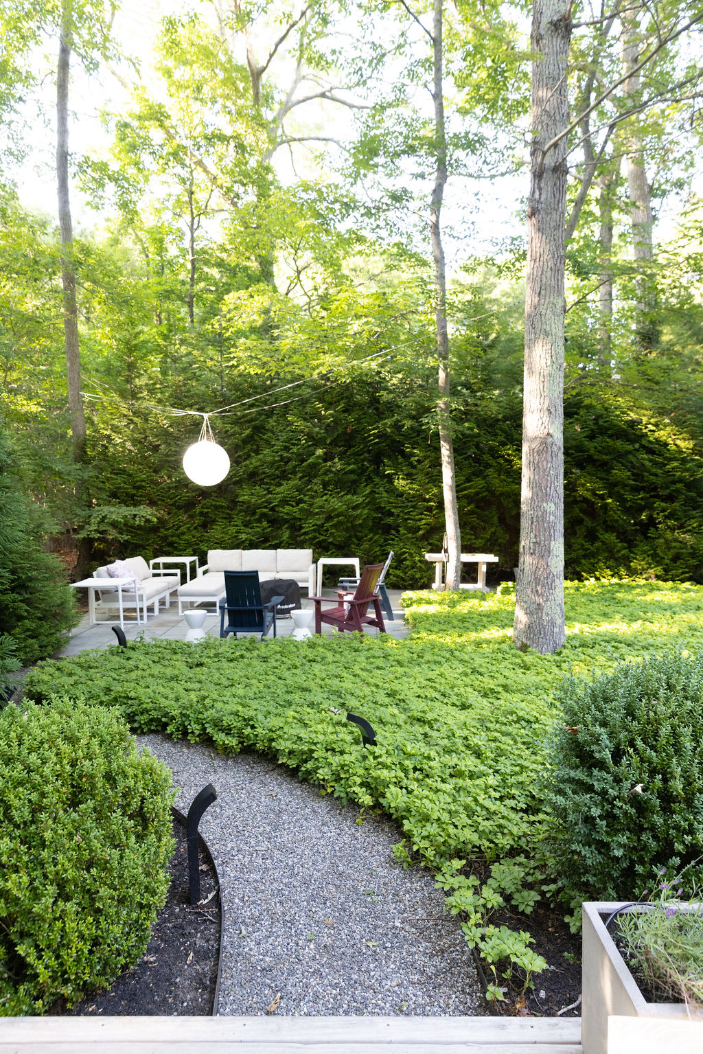 Spacious outdoor lounge area on a light-colored patio, featuring a large sectional sofa, side tables, and colorful Adirondack chairs. A gravel path winds through manicured greenery, leading to the seating area.