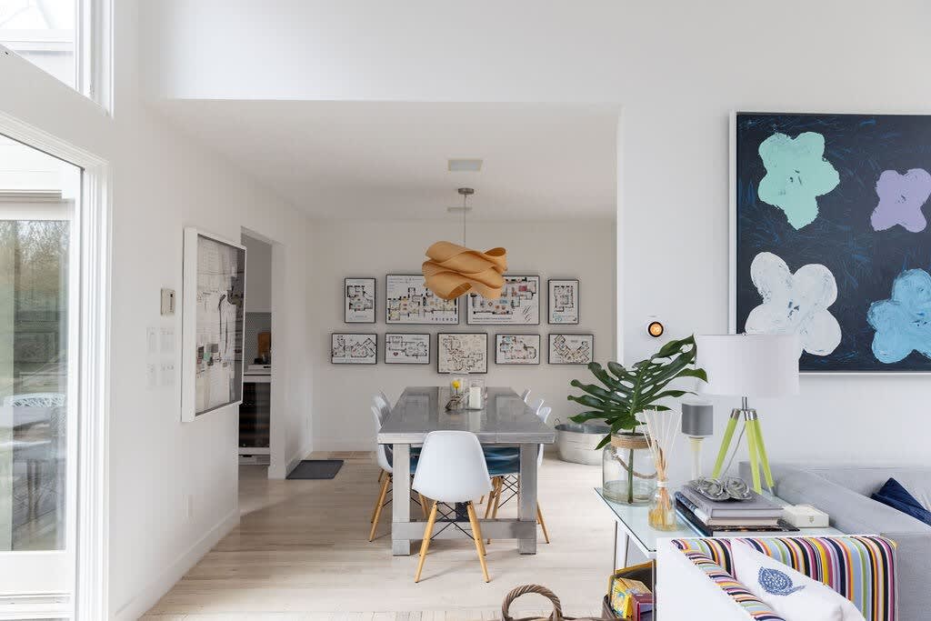 An open-concept view transitions from the living room, with its gray sectional and abstract art, into a bright dining area. The dining room features a long metal table and white chairs, with large windows and a unique wooden pendant light.