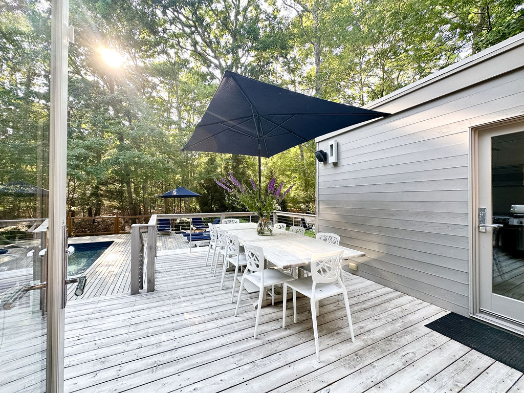Wooden deck with a long white outdoor dining table and matching chairs, shaded by a large black umbrella. The modern gray house exterior is visible, surrounded by dense green trees.