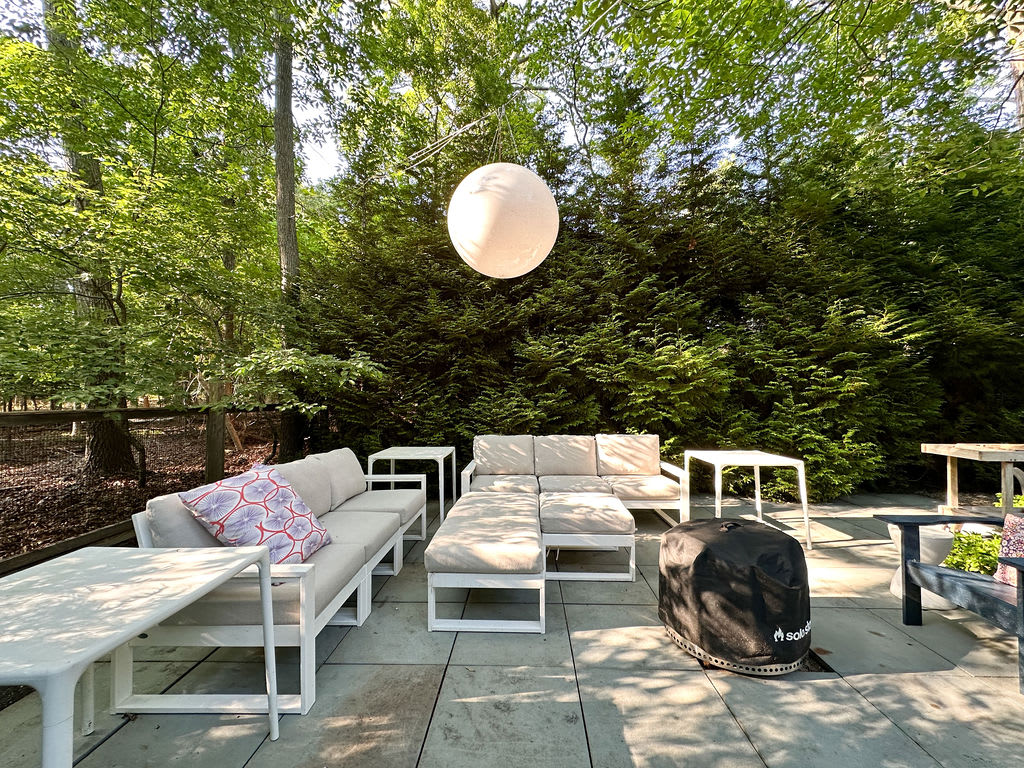 Stone patio featuring a modular outdoor sofa set with light cushions and white frames, along with side tables. A large spherical outdoor light hangs above, and the area is enclosed by a tall green hedge.