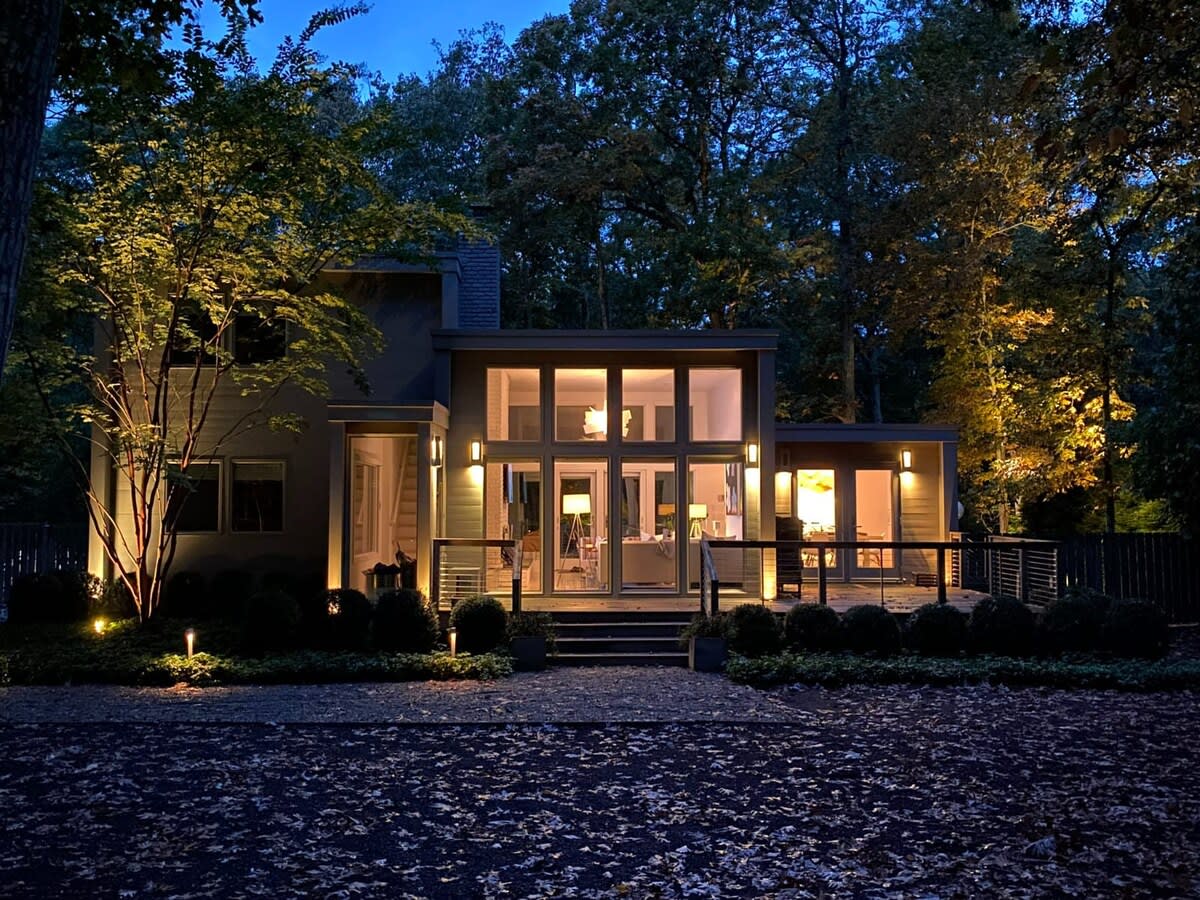 Modern gray house illuminated at night, showcasing its large windows and glass doors, surrounded by trees and subtle landscape lighting. The gravel driveway is covered in fallen leaves, adding to the evening ambiance.