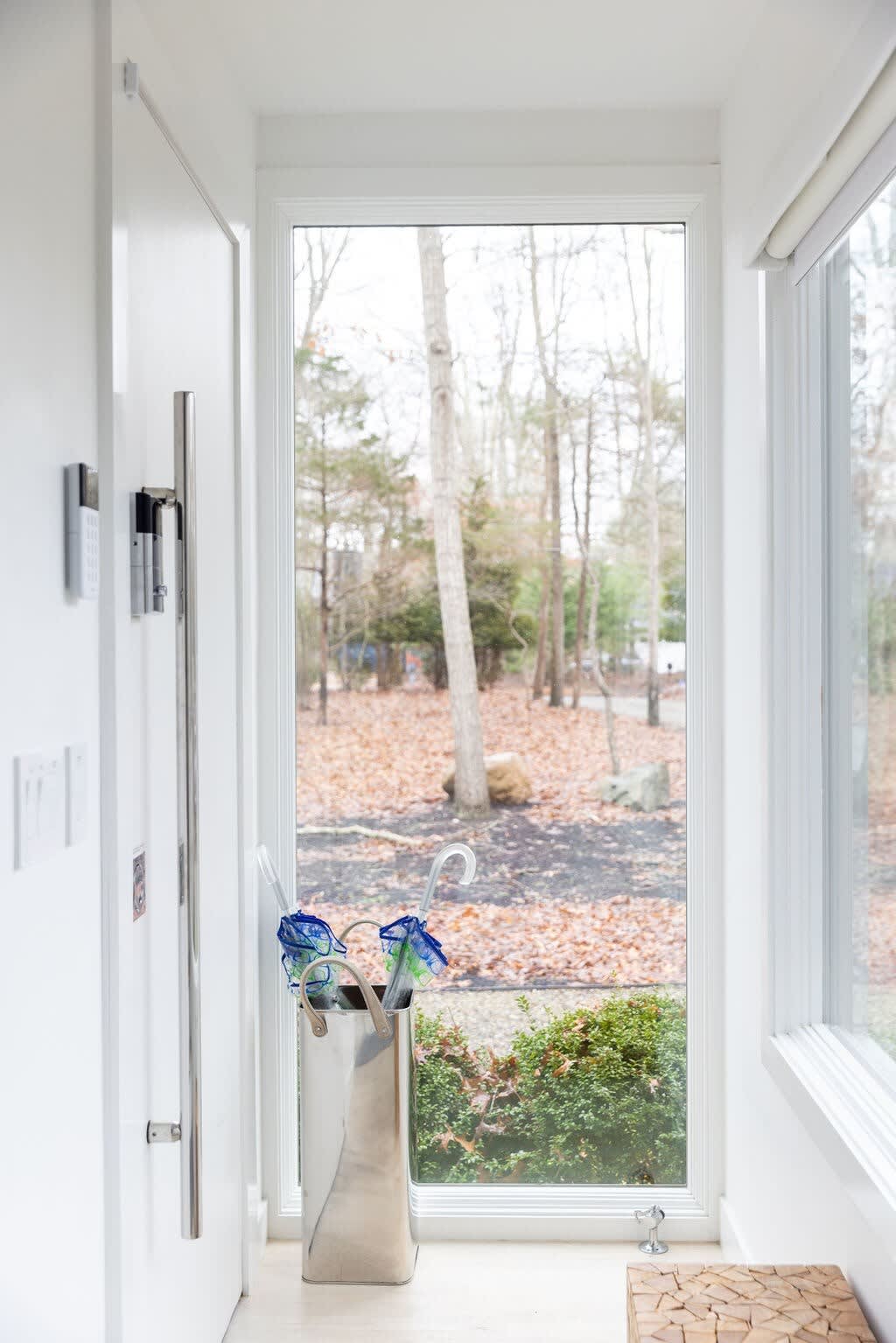 A bright, modern foyer features a tall, narrow window offering a view of a wooded outdoor area with fallen leaves. A sleek umbrella stand with blue umbrellas sits beside the window, next to a white door with a long silver handle and a security panel.