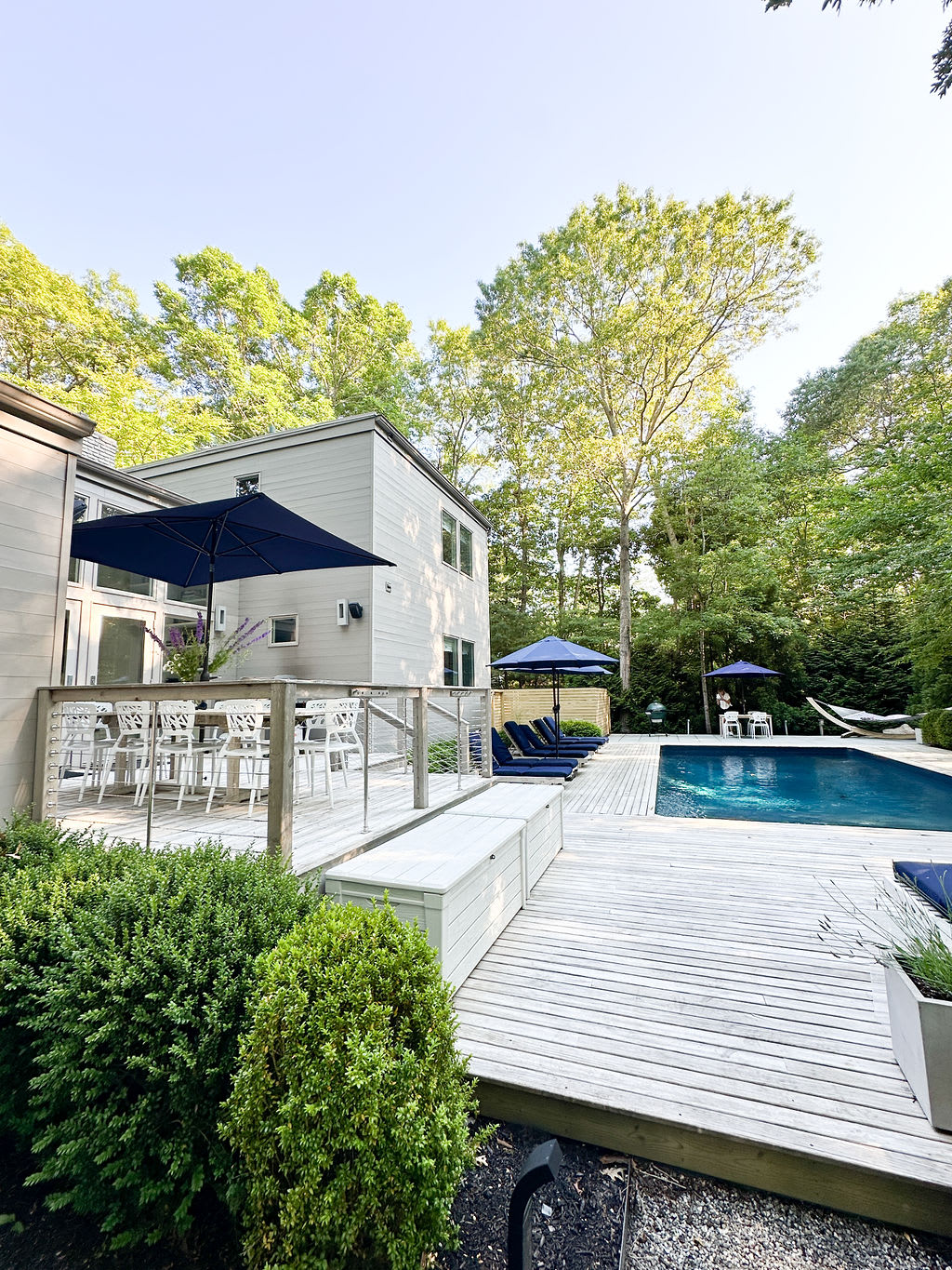 Expansive wooden deck with a rectangular swimming pool, surrounded by lounge chairs with blue cushions and two navy blue umbrellas. The modern house exterior is visible, nestled among tall green trees.