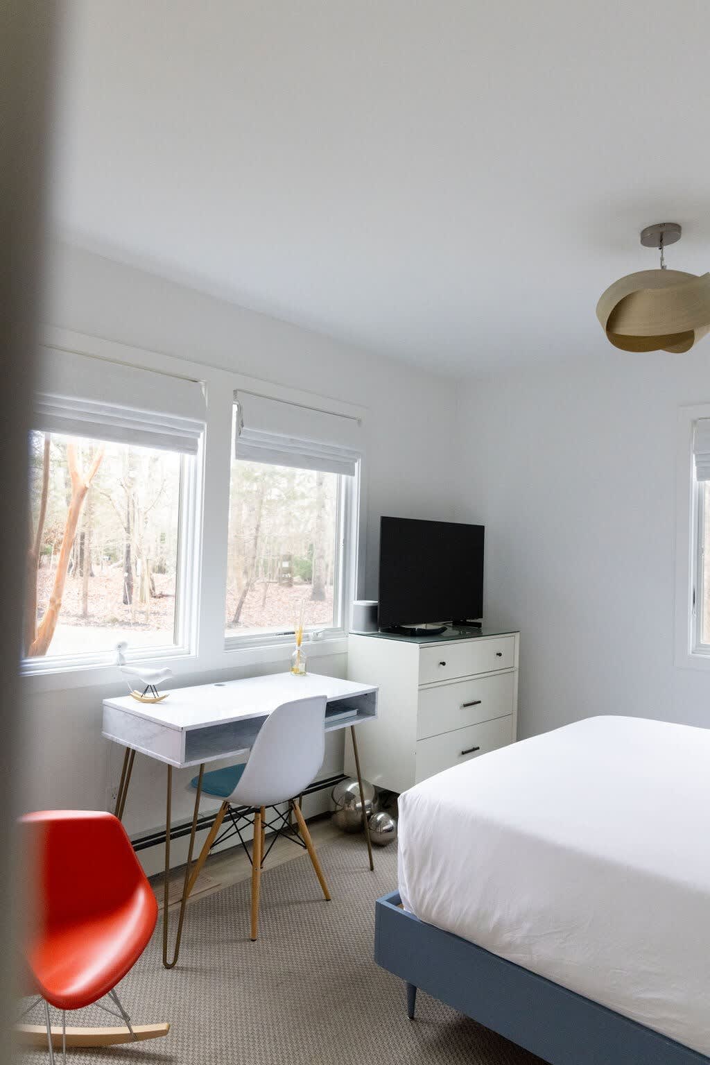 This bright bedroom features a white desk with a modern chair by a large window overlooking trees, and a dresser with a TV. A bed with white linens is partially visible, and an orange rocking chair adds a pop of color to the carpeted floor.