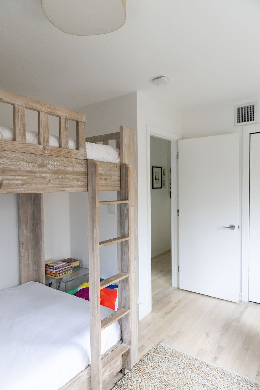 A view of a rustic wooden bunk bed with white bedding and colorful pillows on the lower bunk, set on a light woven rug. The room has light wood floors and white walls, with a doorway leading to another part of the house.