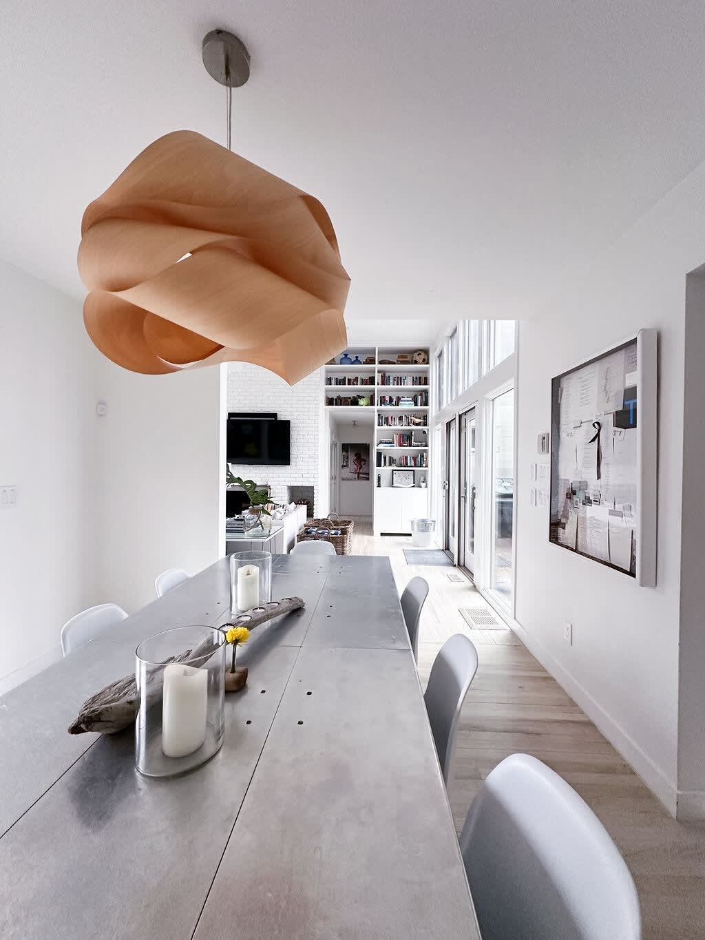 Modern dining room with a long metallic dining table and light gray chairs, illuminated by a unique wooden sculptural pendant light. White walls and light wood floors extend towards a living area with built-in bookshelves.