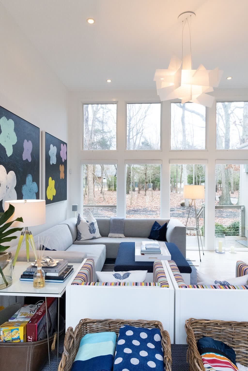 A bright, open-concept living room showcases a gray sectional sofa, a dark blue ottoman coffee table, and two white armchairs with colorful striped cushions. Large windows span the wall, providing abundant natural light and views of the surrounding trees, complemented by modern abstract art.