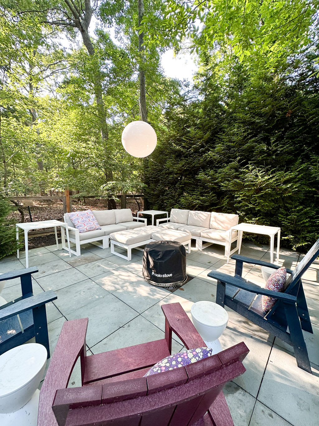 Cozy outdoor lounge area on a paved patio, centered around a black portable fire pit. It features a large L-shaped sectional sofa with light cushions and several colorful Adirondack chairs, all surrounded by lush trees.