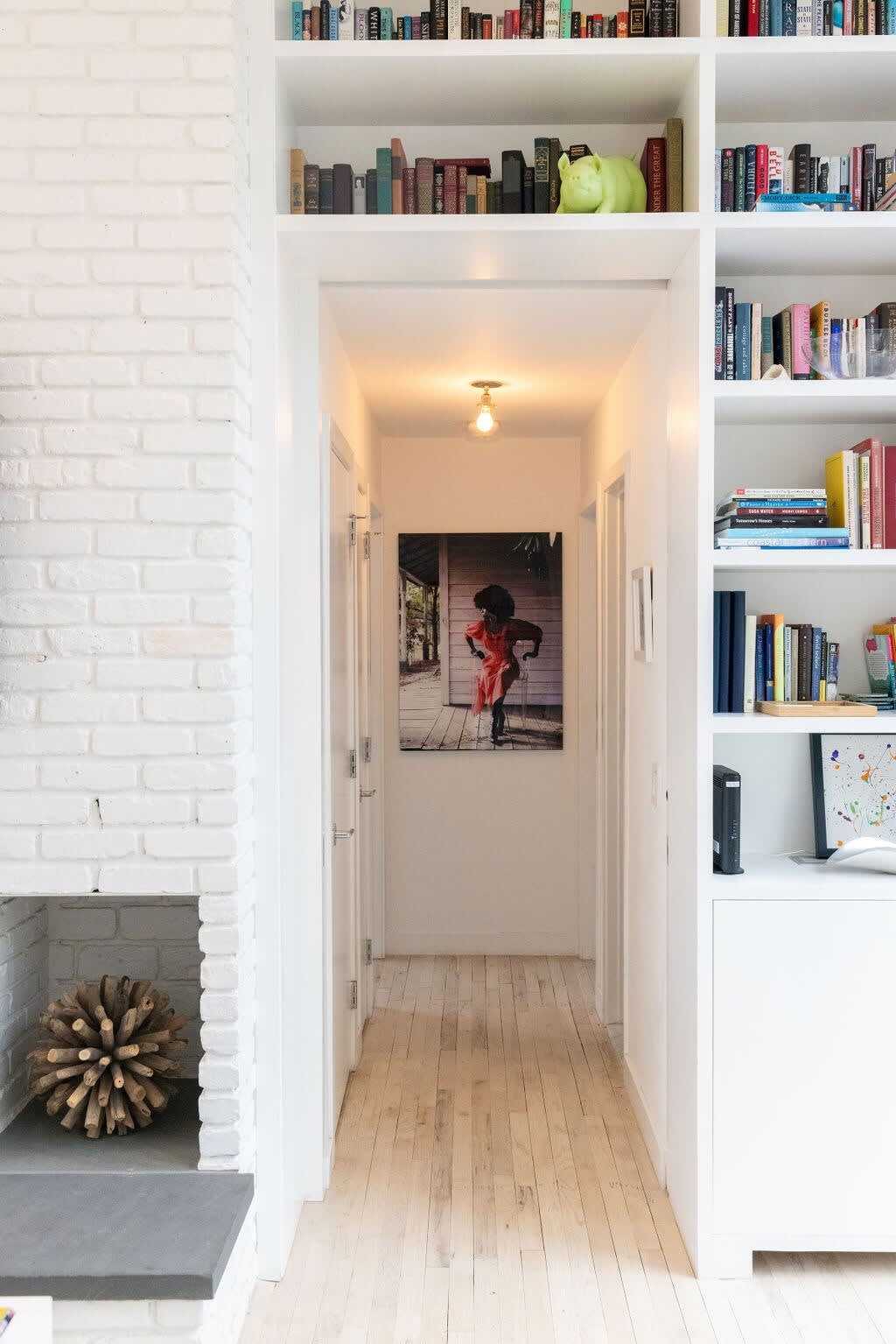 A bright hallway with light hardwood floors leads to a large framed artwork at its end, flanked by white doors. A tall, built-in white bookshelf filled with books and decorative items lines the right wall, next to a white brick fireplace.