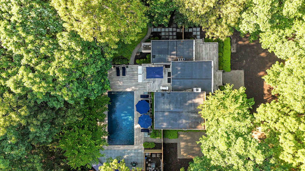 Overhead aerial view of a modern property nestled within a dense forest, showcasing the house with flat roofs, a rectangular swimming pool, and multiple outdoor seating areas. Lush green trees dominate the landscape.