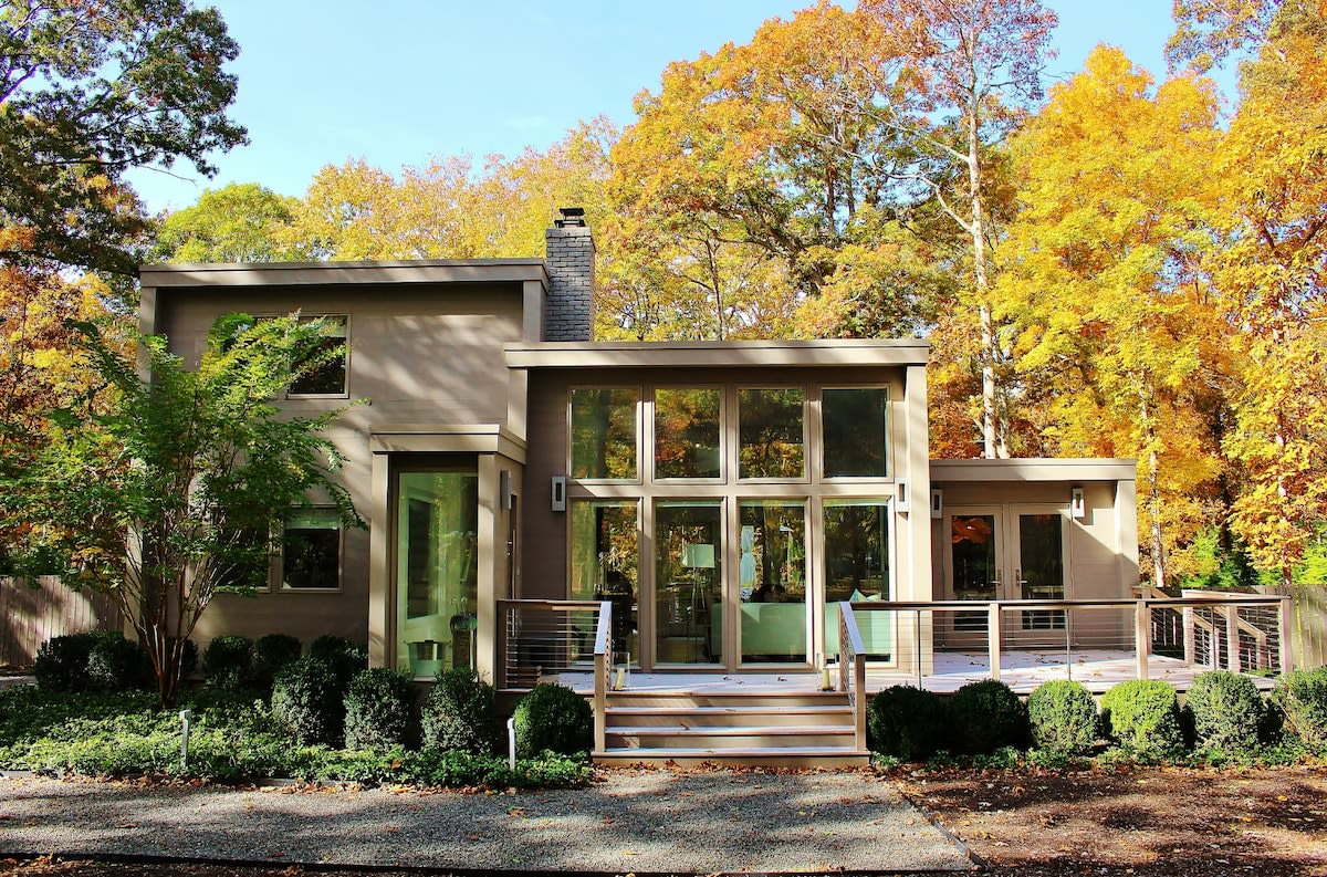 Striking modern house with light gray siding and expansive windows, set against a backdrop of vibrant autumn foliage. A wooden deck with cable railings leads to the front entrance, surrounded by manicured shrubs.