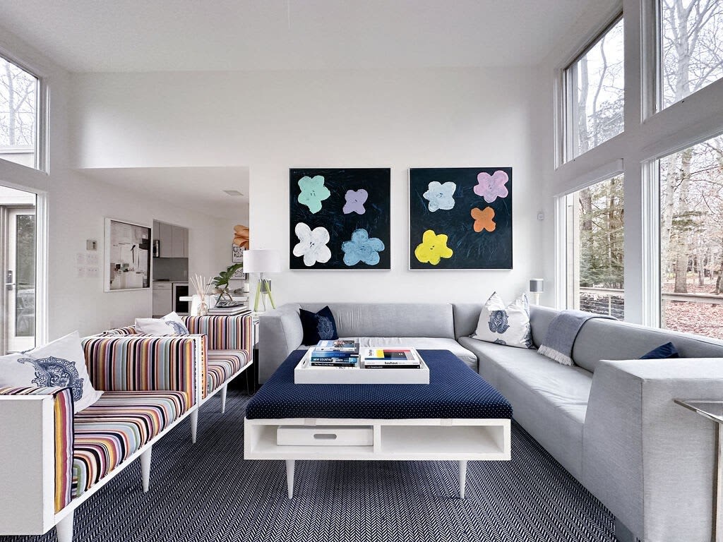 A spacious, modern living room features a large gray sectional sofa and a dark blue upholstered coffee table with books. Two vibrant floral abstract paintings hang above the sofa, and large windows offer views of a wooded outdoor landscape.