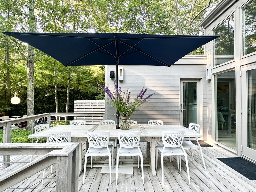 Outdoor dining area on a wooden deck featuring a long white table with eight white chairs, shaded by a large navy blue umbrella. A vase with purple flowers sits on the table, surrounded by lush green trees.
