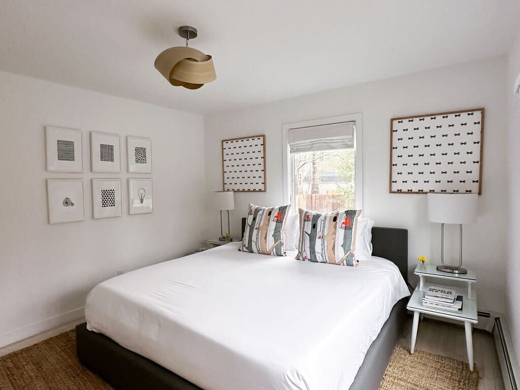 A modern bedroom features a queen-sized bed with white bedding and two decorative pillows, flanked by glass-top nightstands with lamps. The room has white walls, a unique wooden ceiling light fixture, and framed art on the walls.