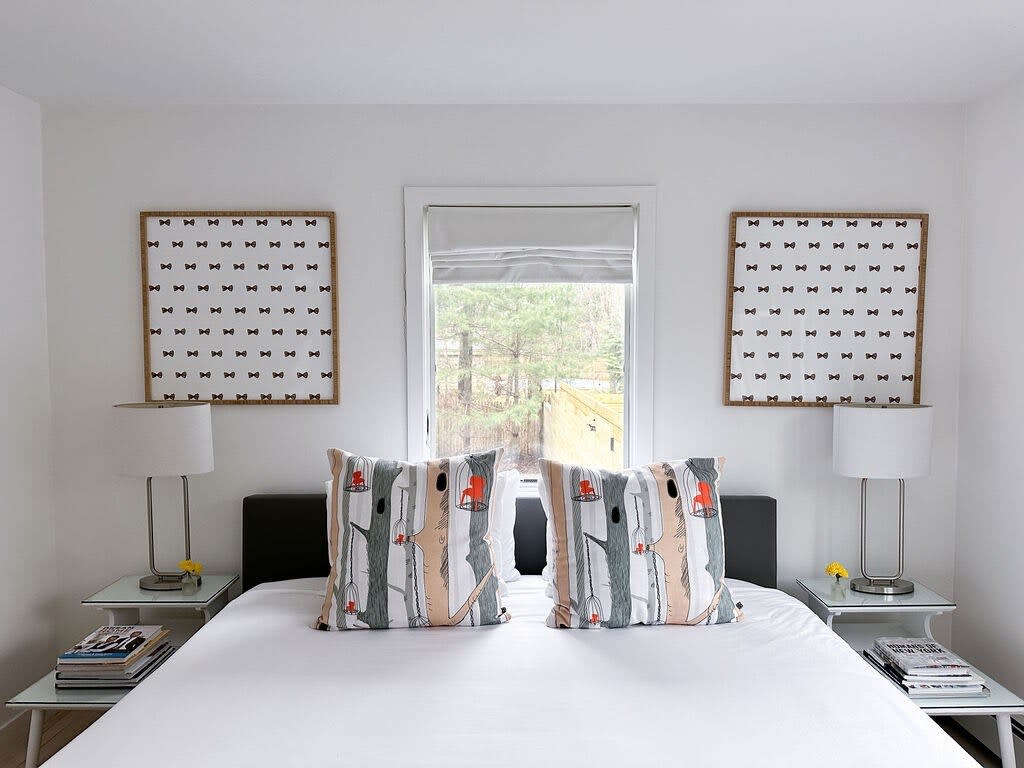 A close-up view of a modern bedroom shows a queen bed with white linens and two patterned pillows, positioned between two glass-top nightstands with lamps. A large window with a roller shade overlooks trees, flanked by two framed art pieces with bow tie patterns.