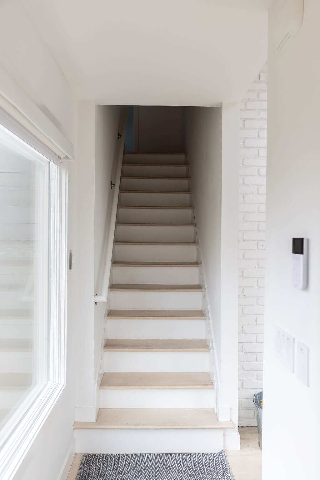 A bright, minimalist interior staircase features light wood treads and white risers, with a simple white handrail leading upwards. A large window on the left provides natural light, and a white brick accent wall is visible on the right.