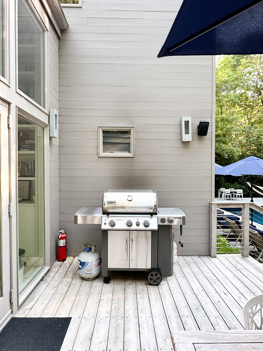 Stainless steel Weber BBQ grill with a propane tank, positioned on a light-colored wooden deck next to the modern gray exterior of the house, with a fire extinguisher nearby.