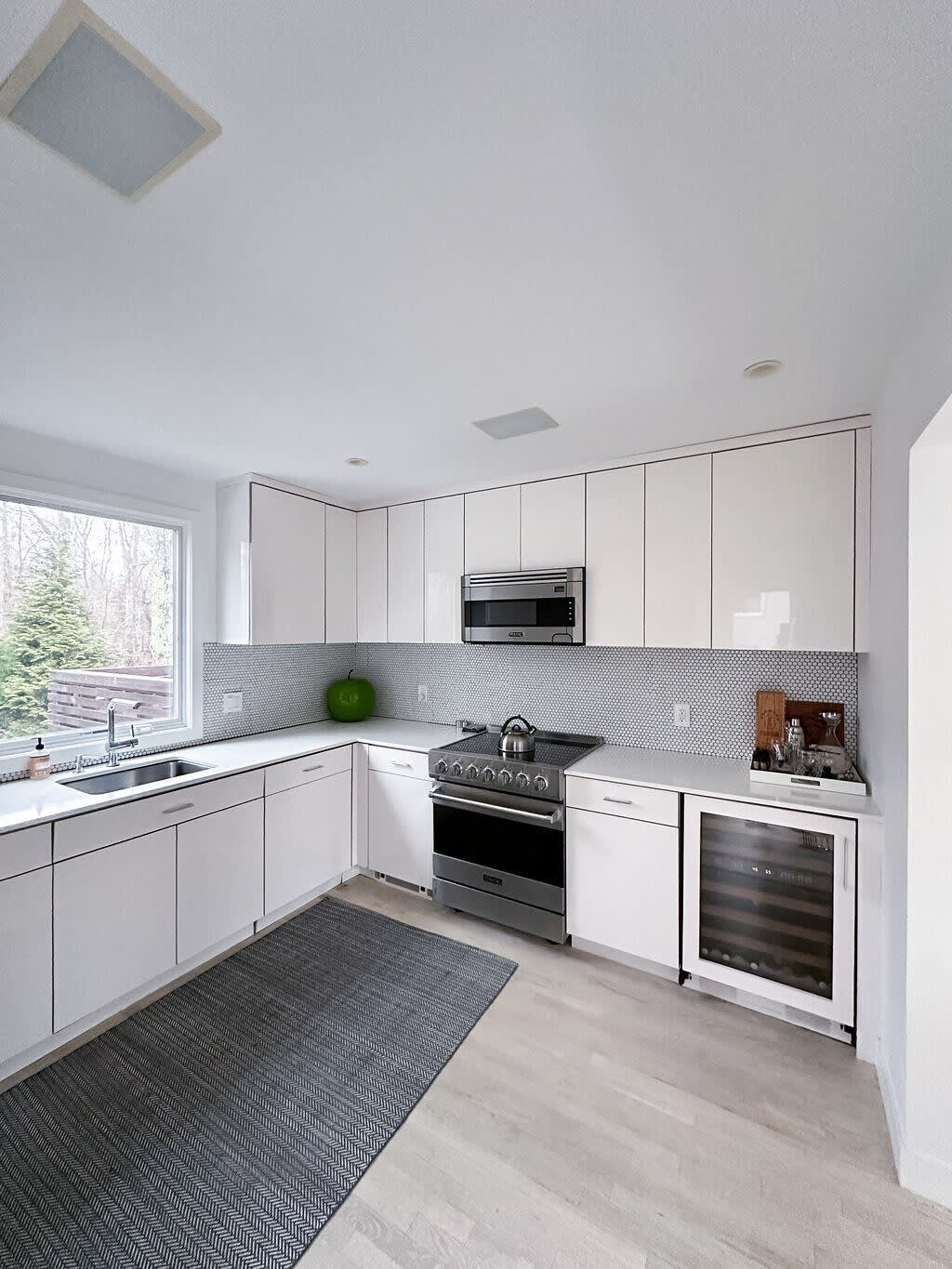 A sleek, modern kitchen features white flat-panel cabinetry, light countertops, and a gray penny tile backsplash. Stainless steel appliances include a Viking range with an overhead microwave, and a built-in wine cooler.