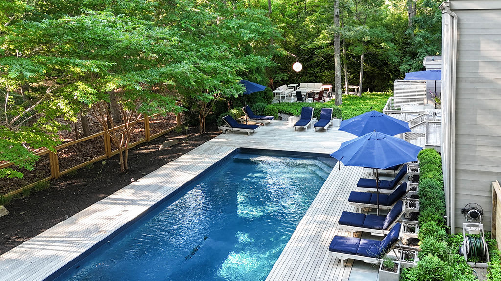 Elevated view of a long rectangular swimming pool surrounded by a light wooden deck and numerous blue lounge chairs with umbrellas. The property is nestled among dense green trees, with a fence visible on one side.