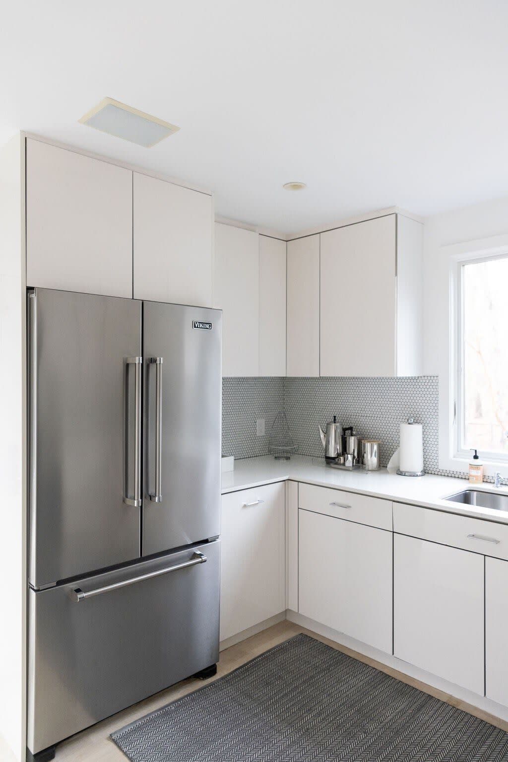 This view of the modern kitchen showcases a large stainless steel French door refrigerator and a sink area with a window overlooking the outdoors. White flat-panel cabinetry and light countertops maintain the minimalist aesthetic, complemented by a gray penny tile backsplash.