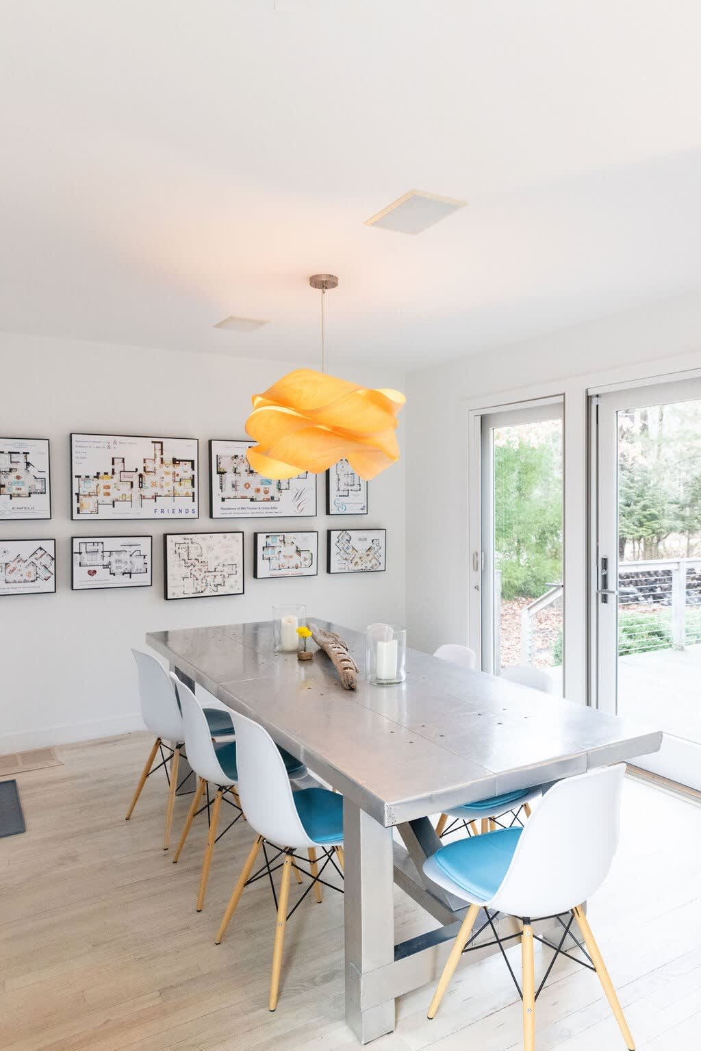 A modern dining room features a long, rectangular metal dining table surrounded by six white chairs with light wood legs and blue seat cushions. A unique, sculptural wooden pendant light hangs above, and a gallery wall of framed floor plans adorns the back wall.