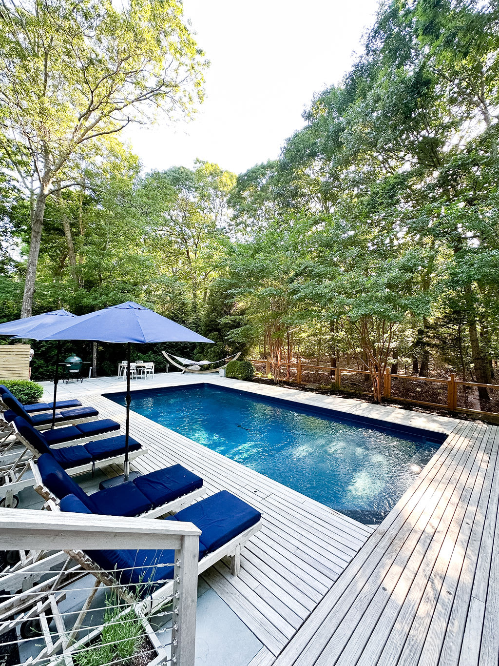 Inviting outdoor swimming pool surrounded by a light-colored wooden deck, multiple blue lounge chairs with matching umbrellas, and lush green trees providing natural privacy.