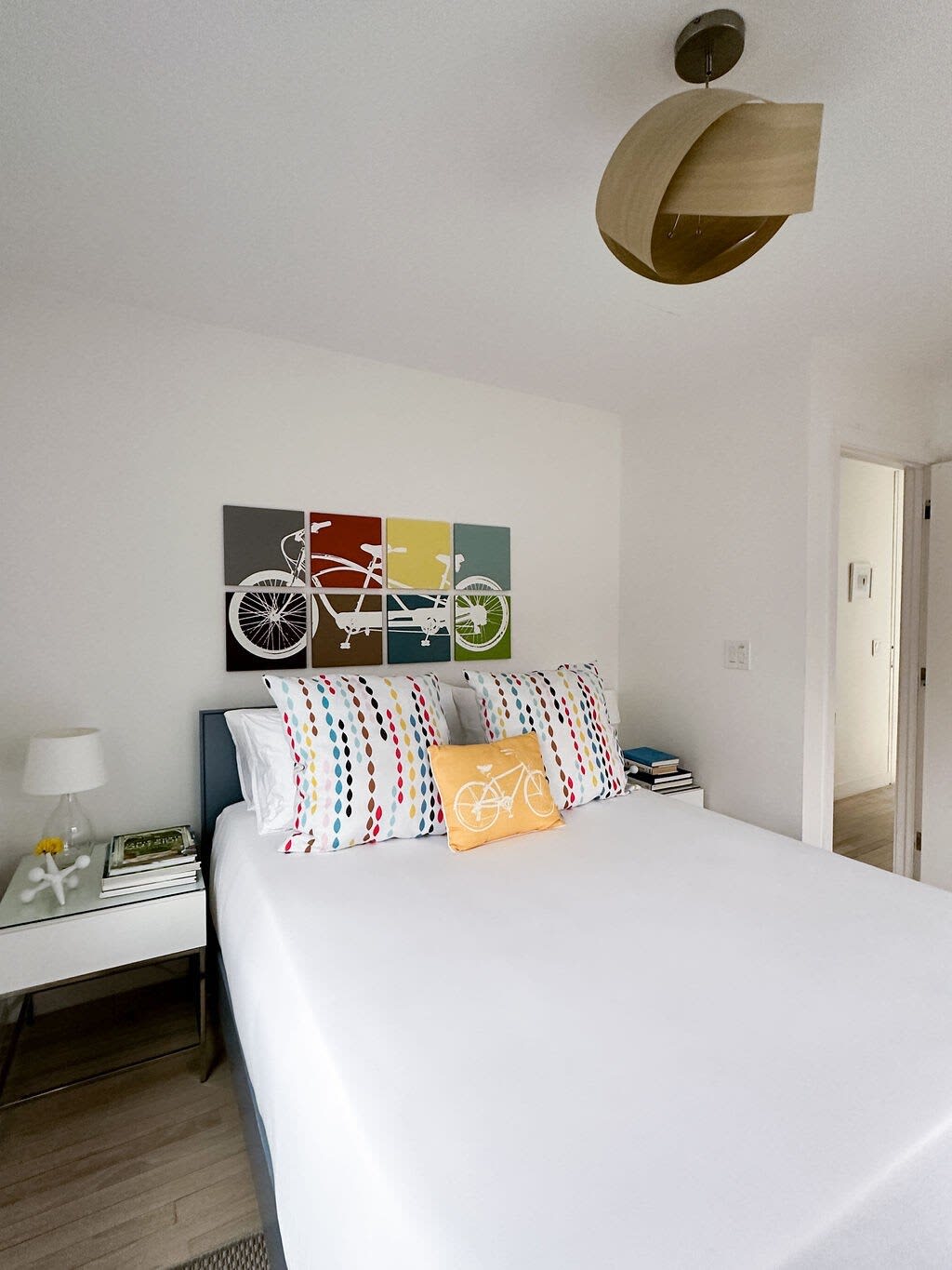 A colorful bedroom showcases a bed with white linens and bright, patterned pillows, complemented by a four-panel bicycle art piece above the headboard. A modern wooden ceiling light fixture hangs overhead, and a glass-top nightstand with a lamp is visible beside the bed.
