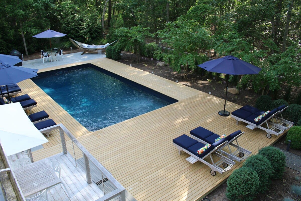 Overhead view of a rectangular swimming pool with a dark blue liner, surrounded by an expansive light-colored wooden deck furnished with lounge chairs and umbrellas, nestled within a lush green wooded area.