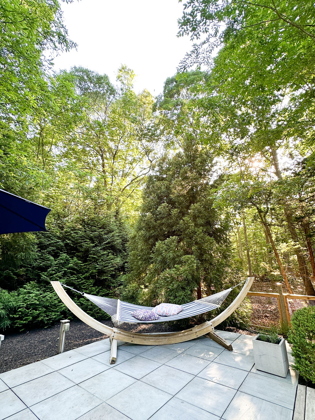 Relaxing hammock with striped cushions on a light-colored tiled patio, supported by a curved wooden stand. Lush green trees and foliage create a private, serene outdoor setting.