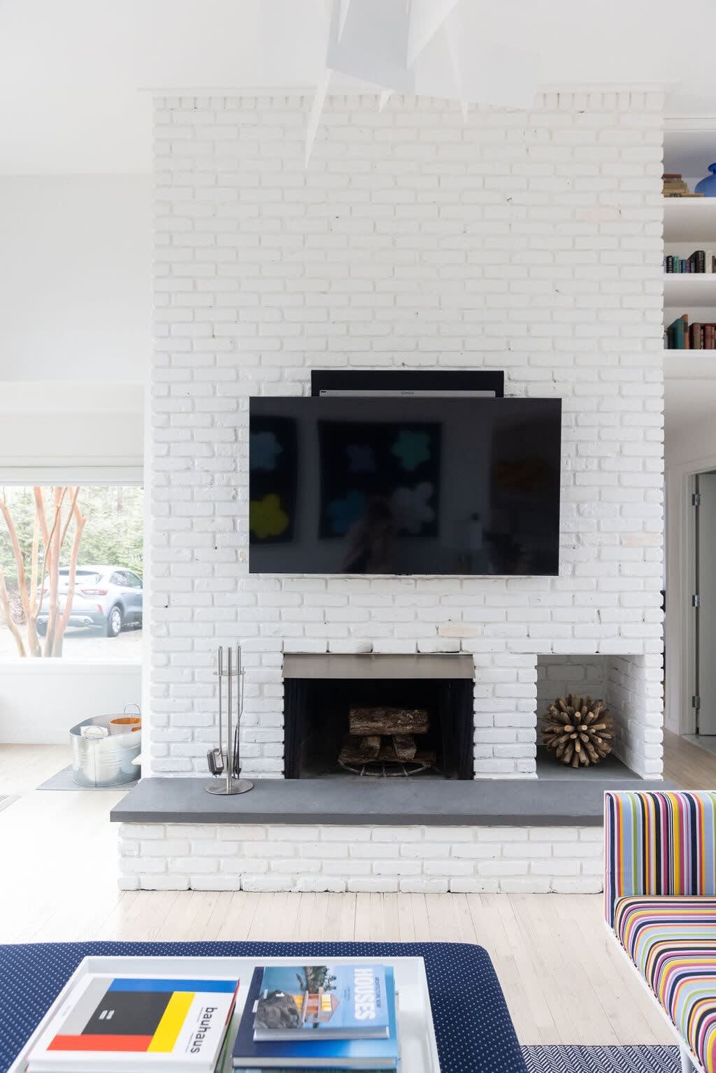 A modern living room features a prominent white painted brick fireplace with a flat-screen TV mounted above it. A dark gray hearth extends across the base, with a firebox and decorative wooden elements in built-in niches.