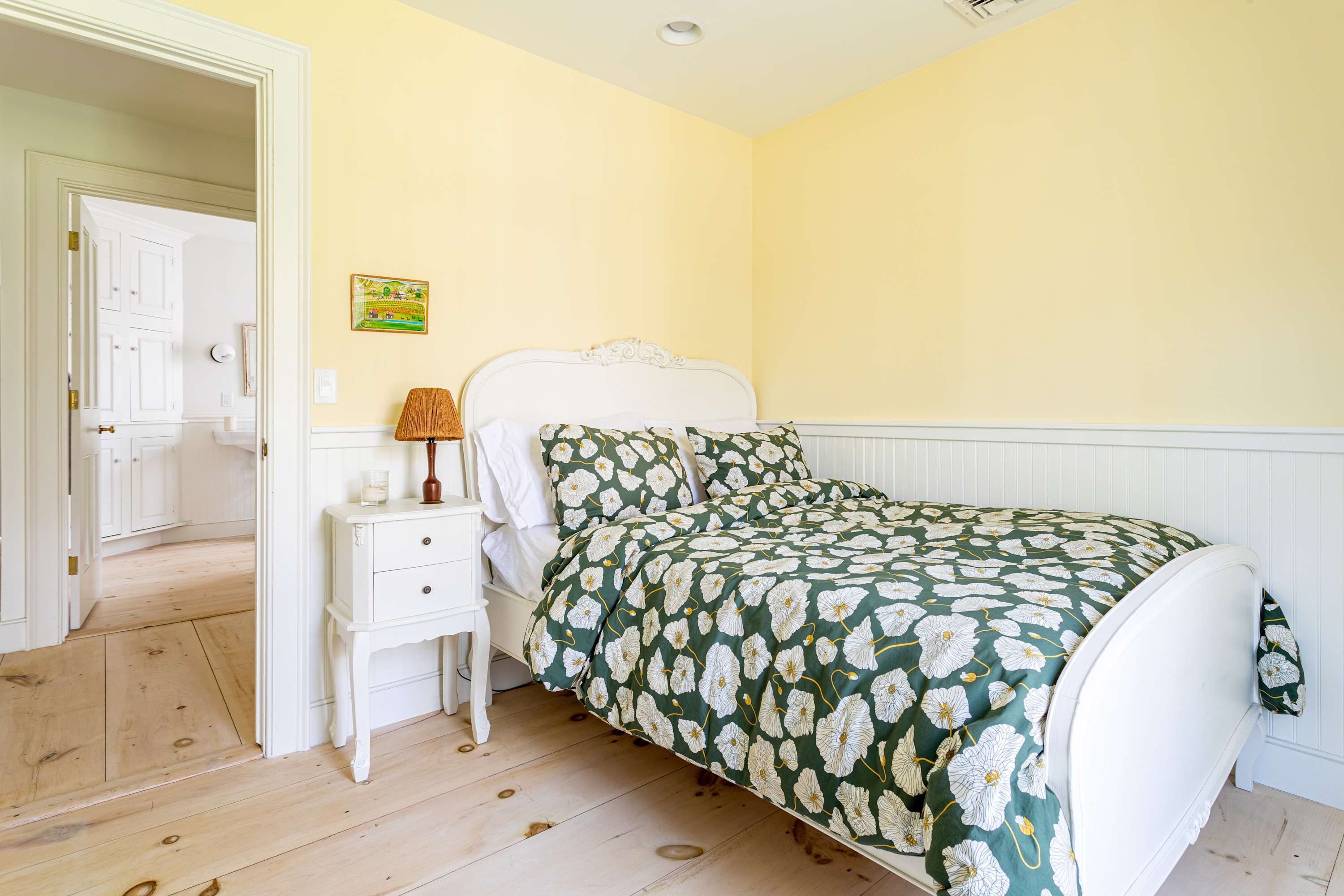 View of the cheerful bedroom with pale yellow walls and light wood floors, showing the white bed with dark green floral bedding and a white nightstand with a lamp. A doorway leads to an adjacent bathroom.
