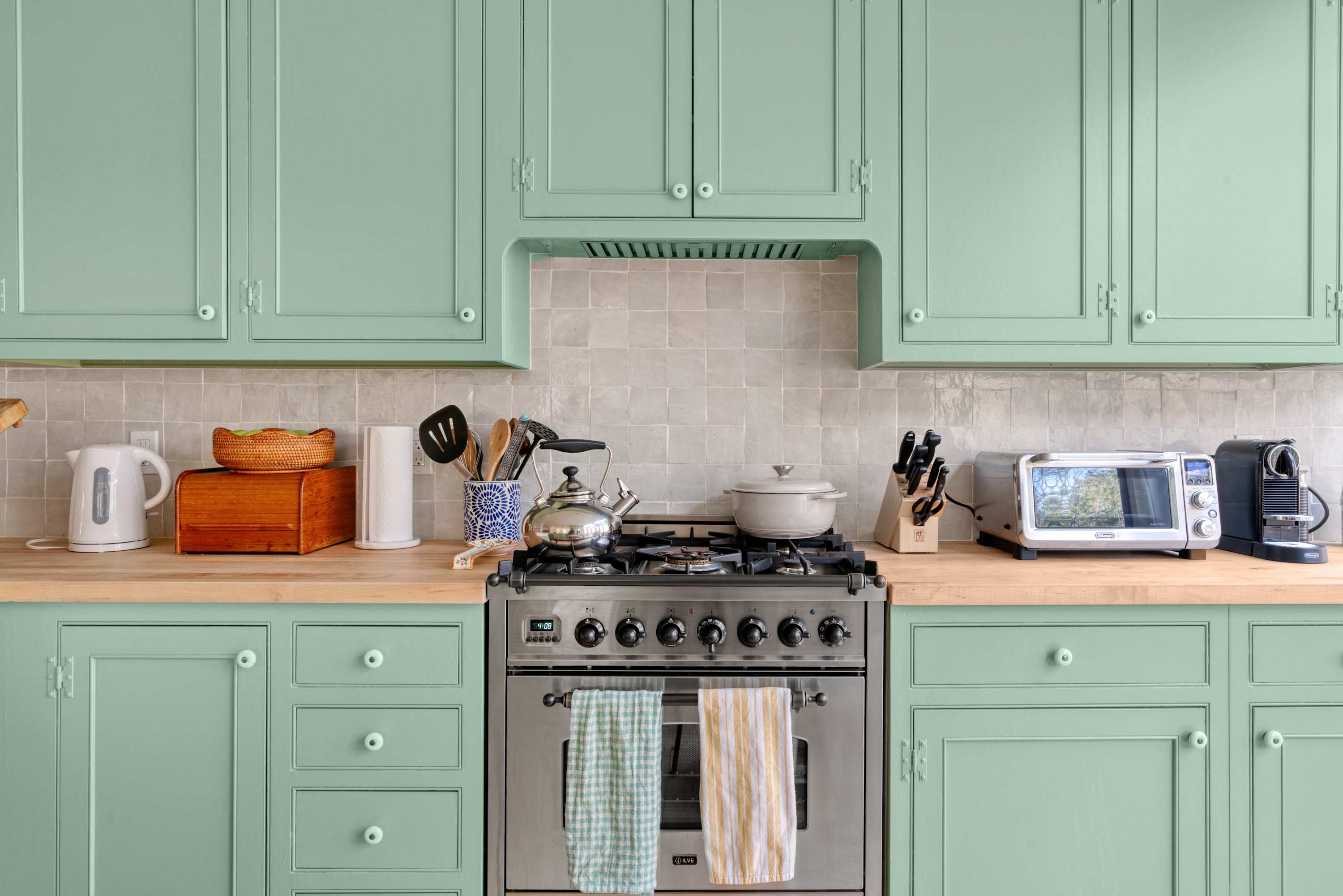 A well-appointed kitchen showcases light green shaker-style cabinets, light wood countertops, and a stainless steel range with a coordinating hood. A subtle light gray tiled backsplash adds texture, while various modern kitchen appliances and utensils are neatly arranged.