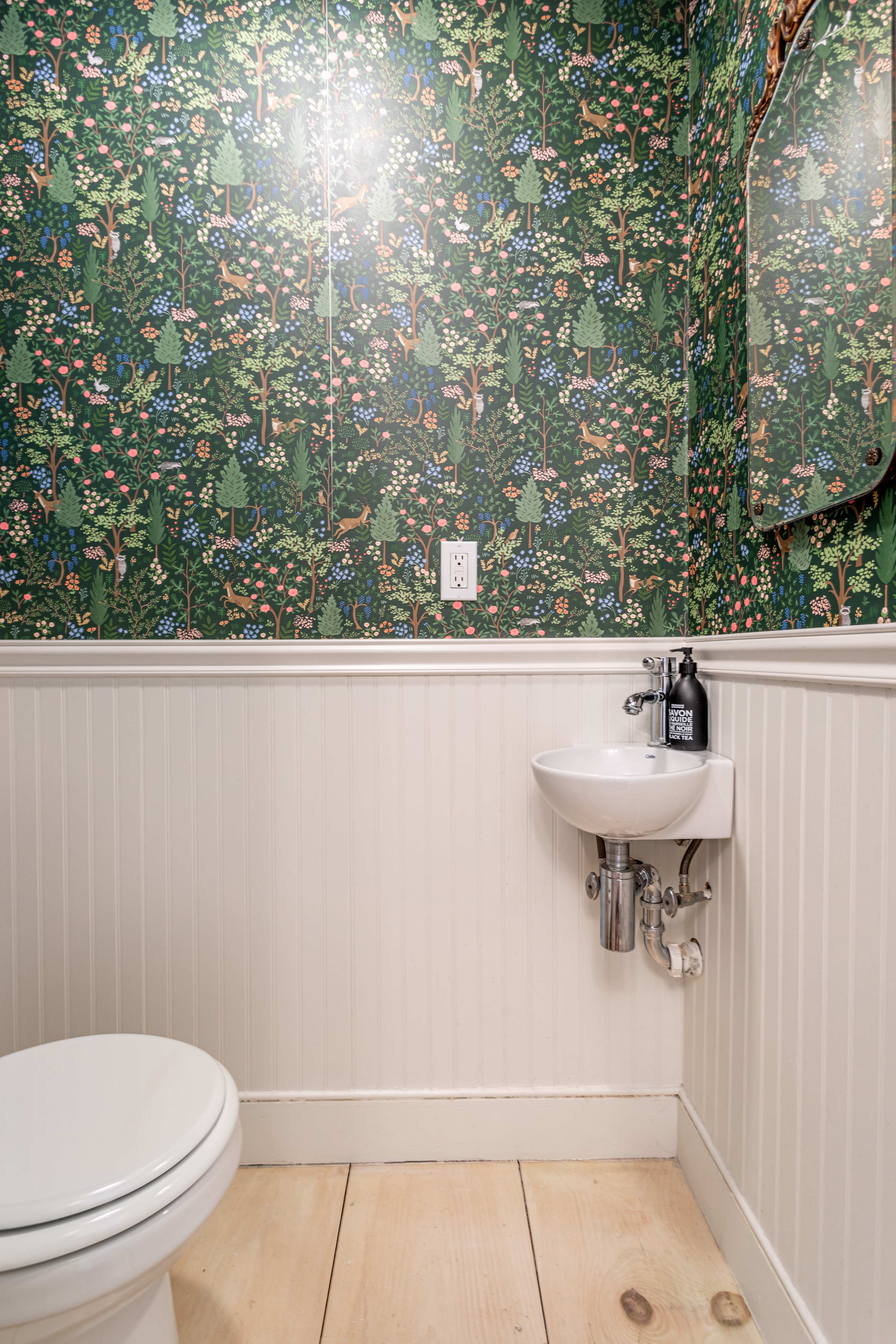 A charming powder room featuring a white toilet and a small wall-mounted sink, with a vibrant green forest-themed wallpaper above white beadboard wainscoting.