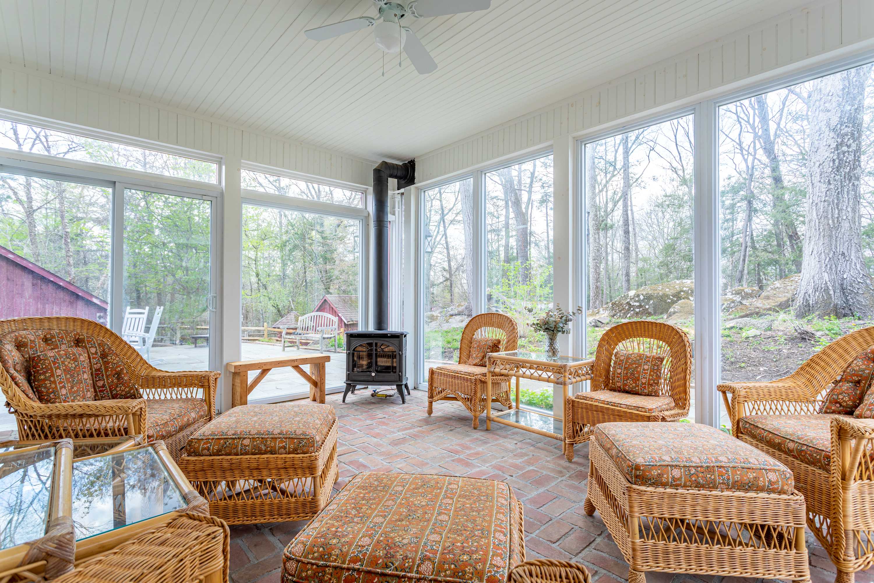 A sun-drenched enclosed porch with a brick floor, furnished with comfortable wicker chairs and ottomans. Large windows offer views of the surrounding trees, and a black wood-burning stove provides warmth.
