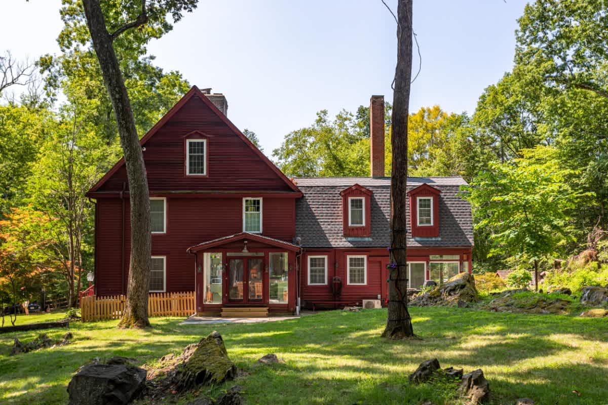 Grand red-sided house with multiple windows and dormers, set amidst a lush green lawn and towering mature trees, featuring a covered entrance and a small wooden fence.