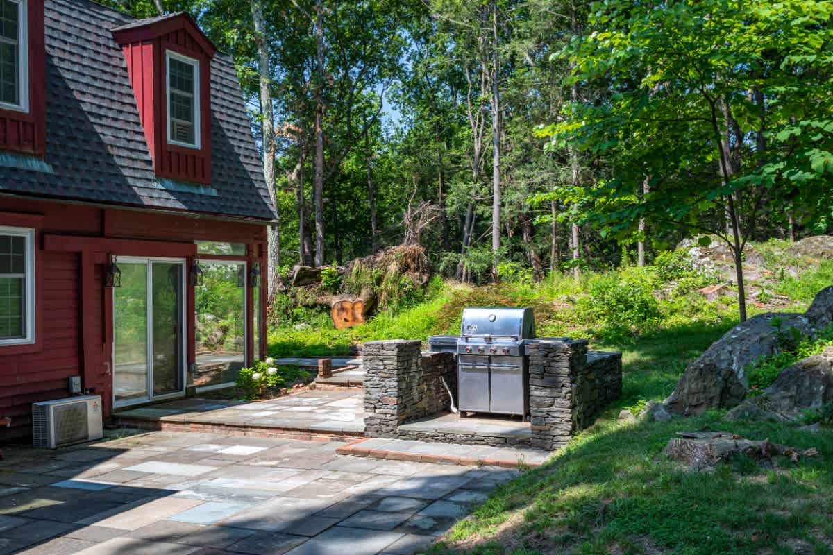 Stainless steel BBQ grill integrated into a stone-built counter on a flagstone patio, adjacent to the red house, with a natural, rocky, and wooded landscape in the background.